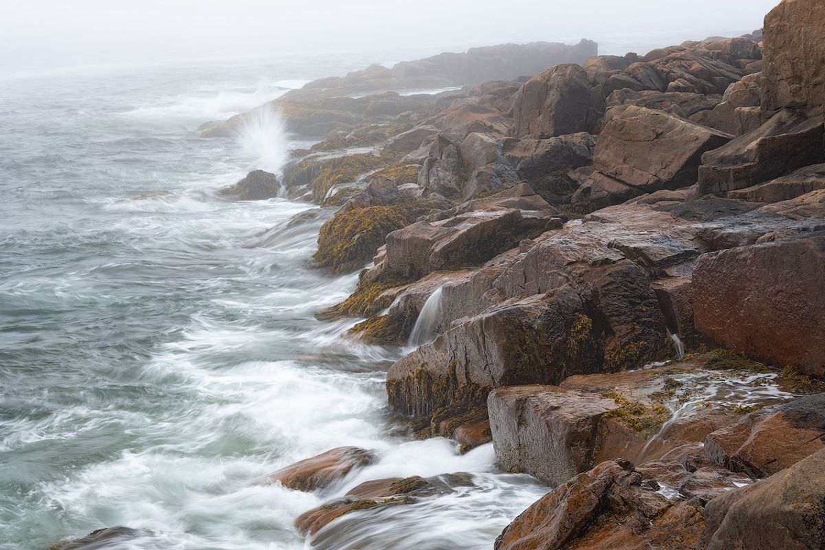 Dramatic waves crash against rugged rocky coastline with ocean spray and fog in the distance.