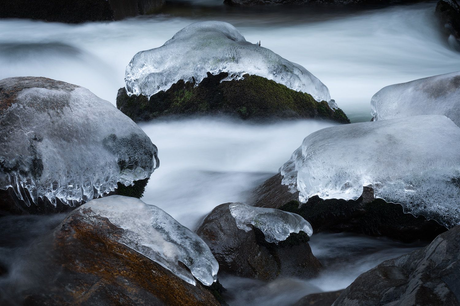 Ice formations cling to dark rocks in a frozen stream creating dramatic winter patterns.