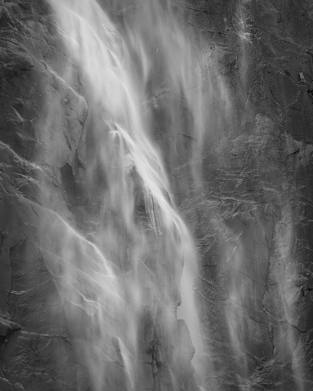 Misty waterfall cascades down dark rocky cliff face in dramatic black and white.