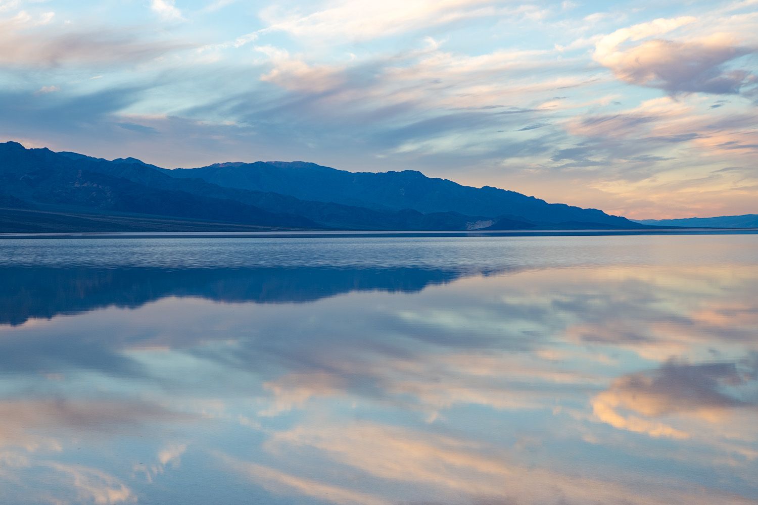Mountain silhouette reflects perfectly in calm lake waters during pastel sunrise in Death Valley.