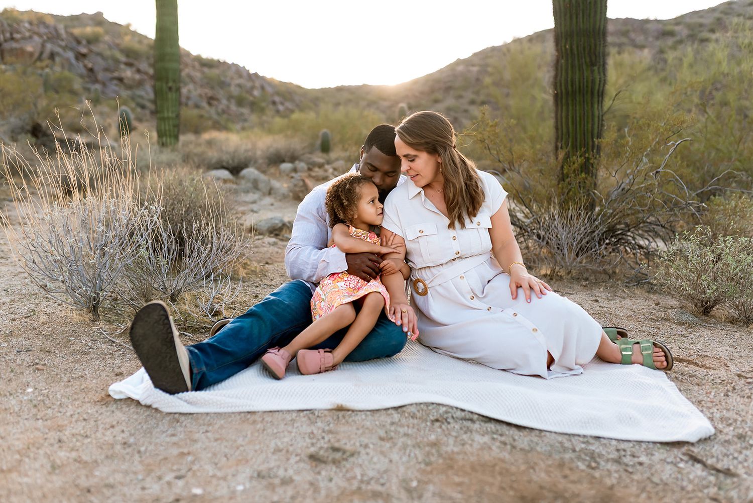 A loving family sits together on a white blanket in a desert landscape with saguaro cacti and mountains in the background.