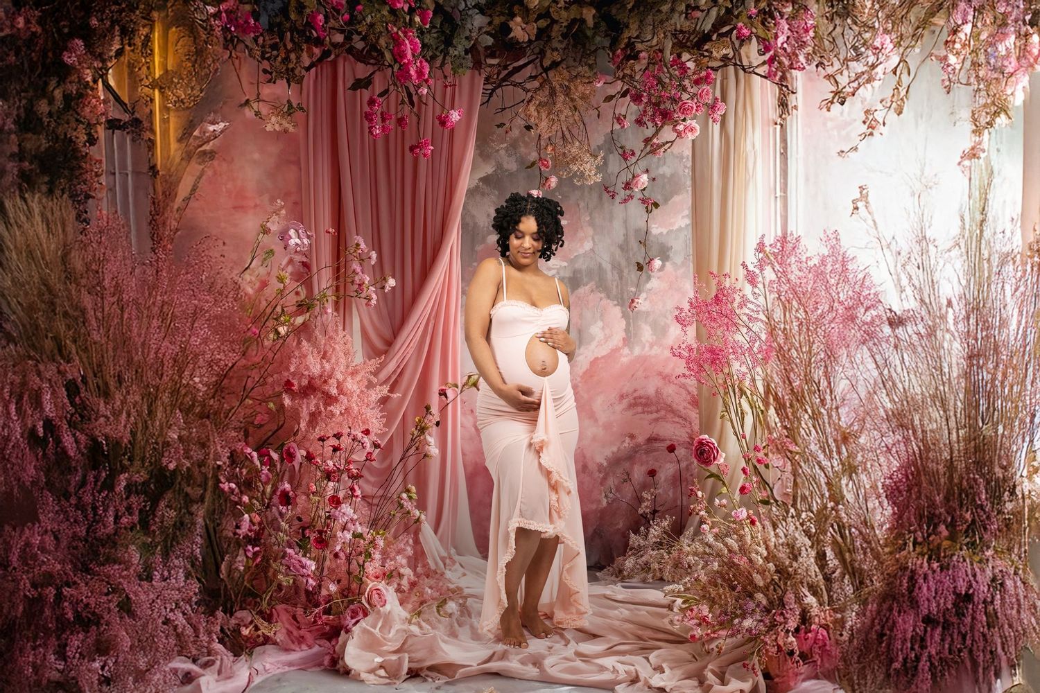 Maternity portrait of woman in pink gown surrounded by florals and drapery in styled NYC studio setup.