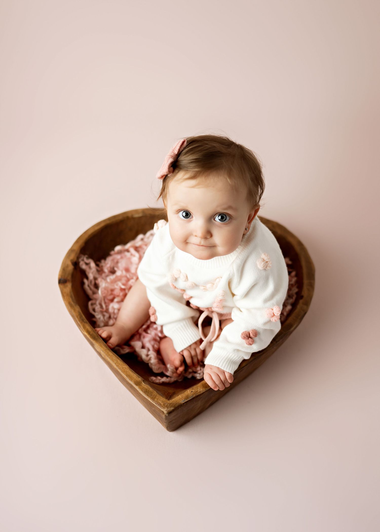 Sleeping baby wearing white outfit rests in heart-shaped wooden bowl with pink textured blanket on neutral background.