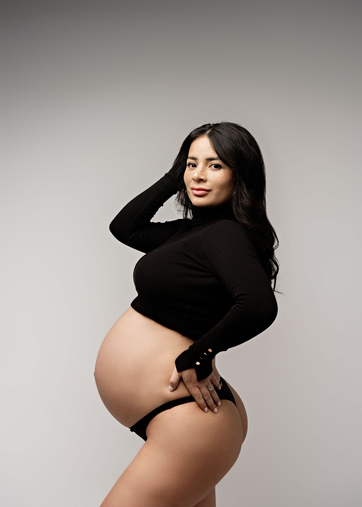 Side view of an elegant maternity photoshoot featuring a woman in black against a neutral gray background.