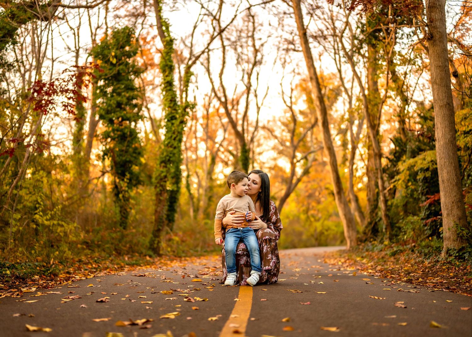 A quiet paved path through autumn woods with fall foliage and warm golden sunlight filtering through the trees.