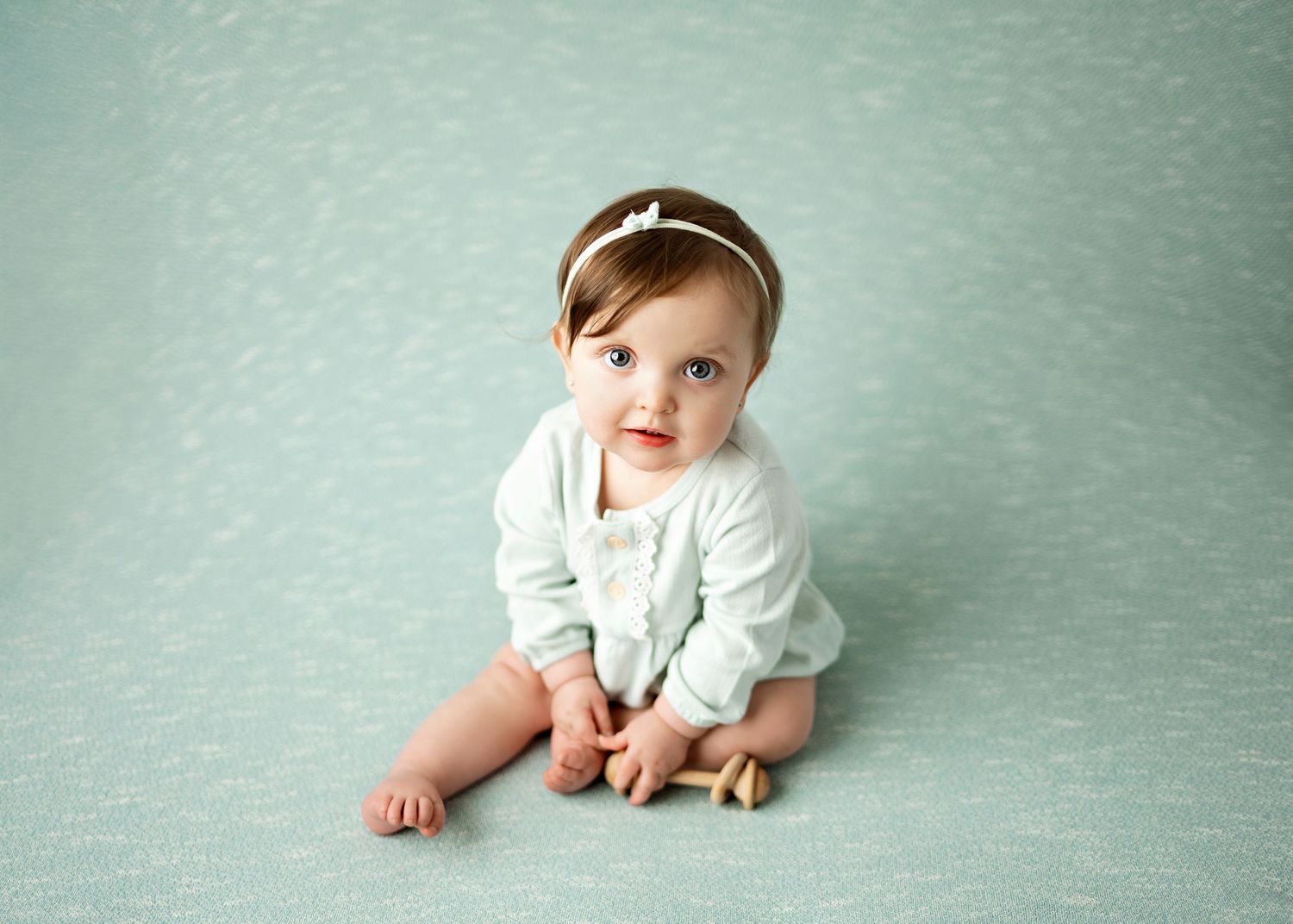 Baby in white outfit sits on minty blue backdrop wearing a headband.