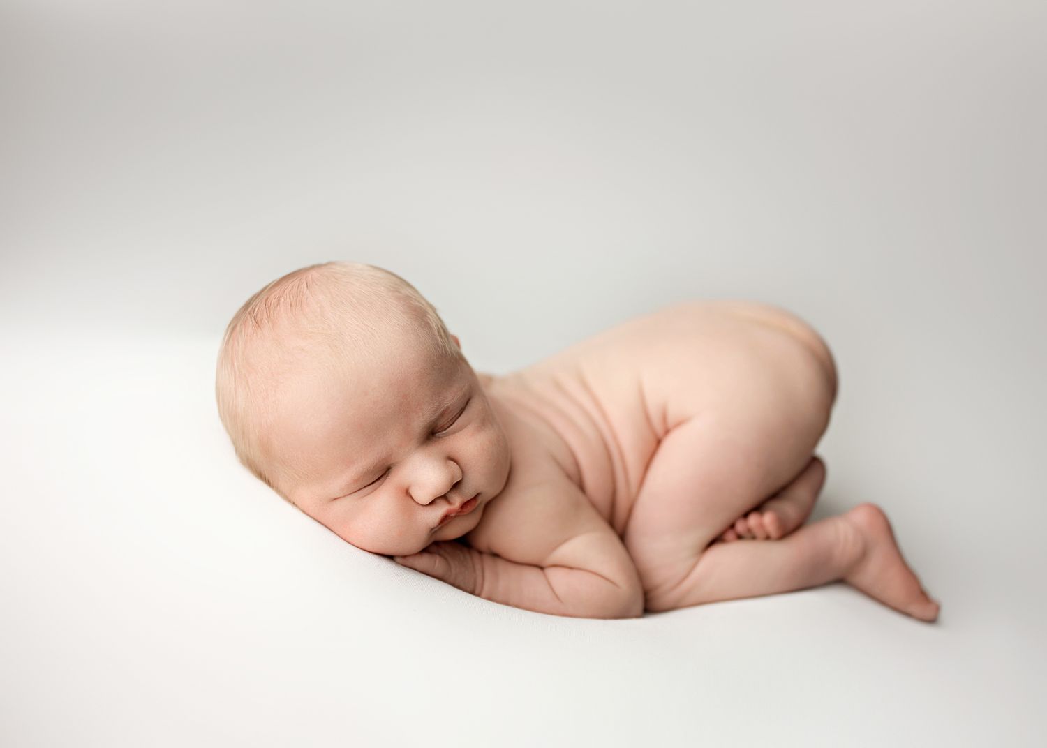 A peaceful sleeping newborn baby curled up in fetal position on soft white backdrop during professional photo session.