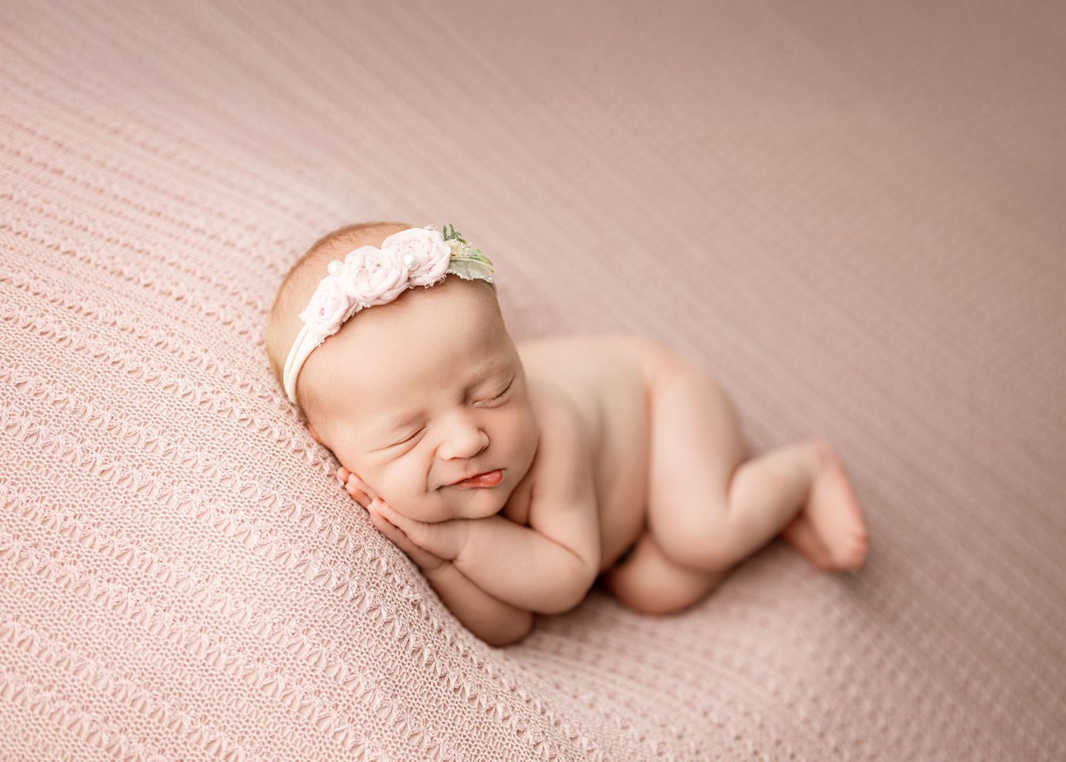 Peaceful sleeping newborn baby wearing delicate white headband resting on soft pink textured blanket.