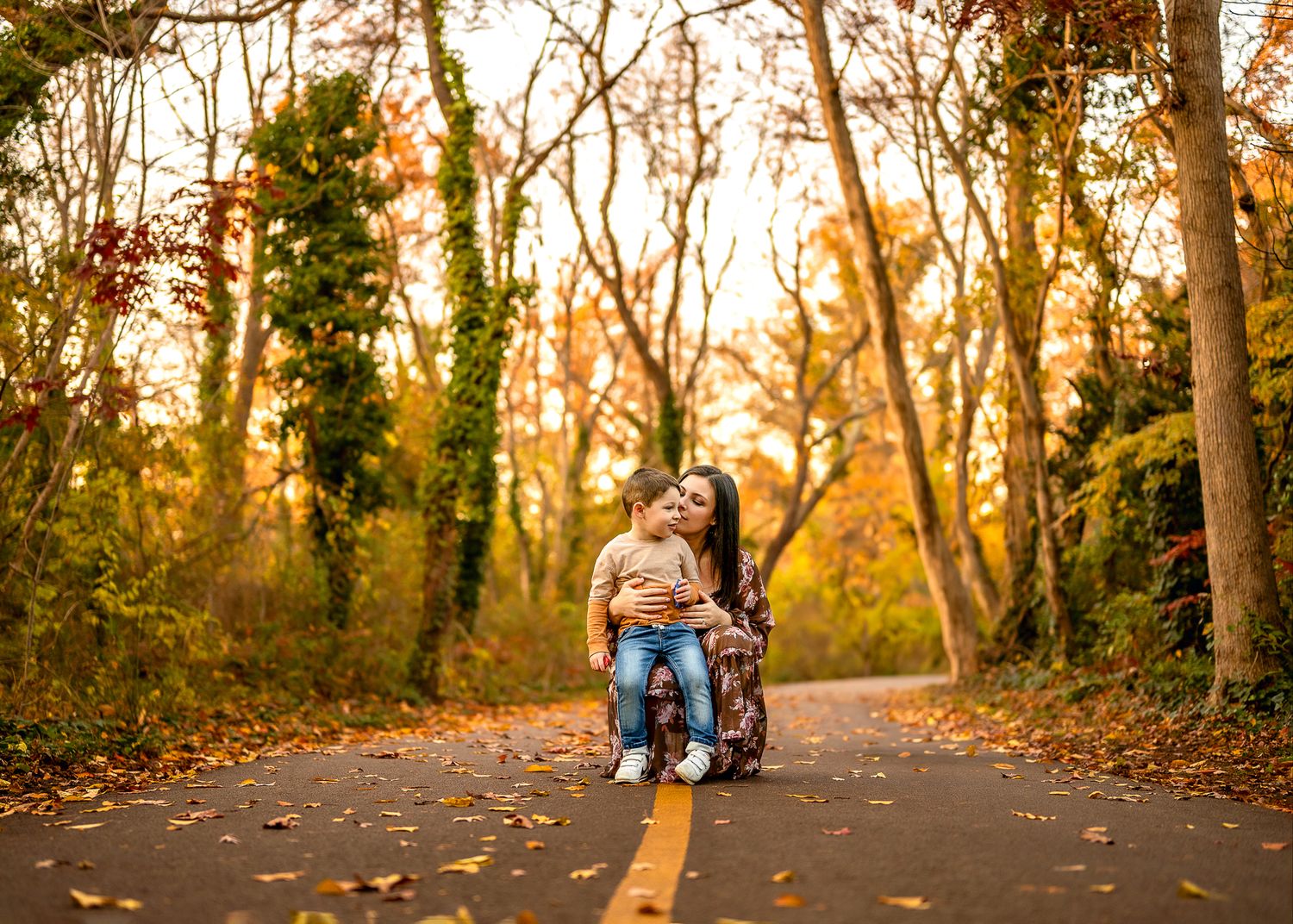 Two people sitting together on a skateboard on a tree-lined path during golden autumn sunset.