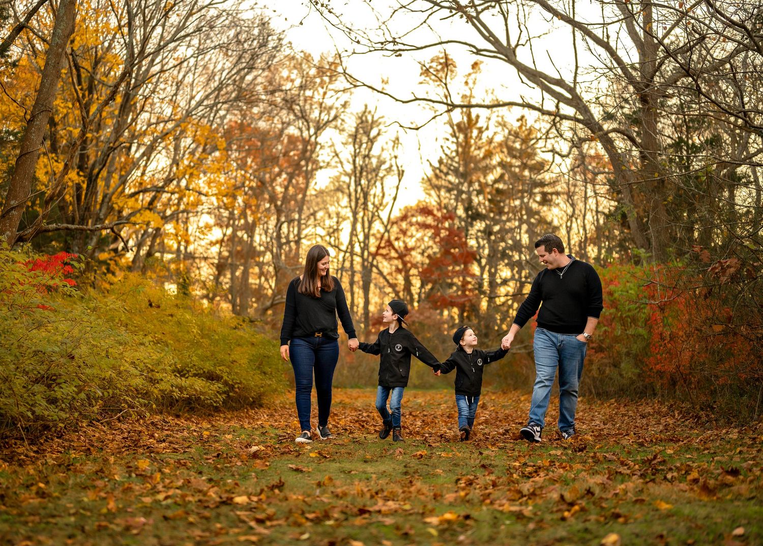 A group walks together down a leaf-covered autumn trail surrounded by golden trees at sunset.