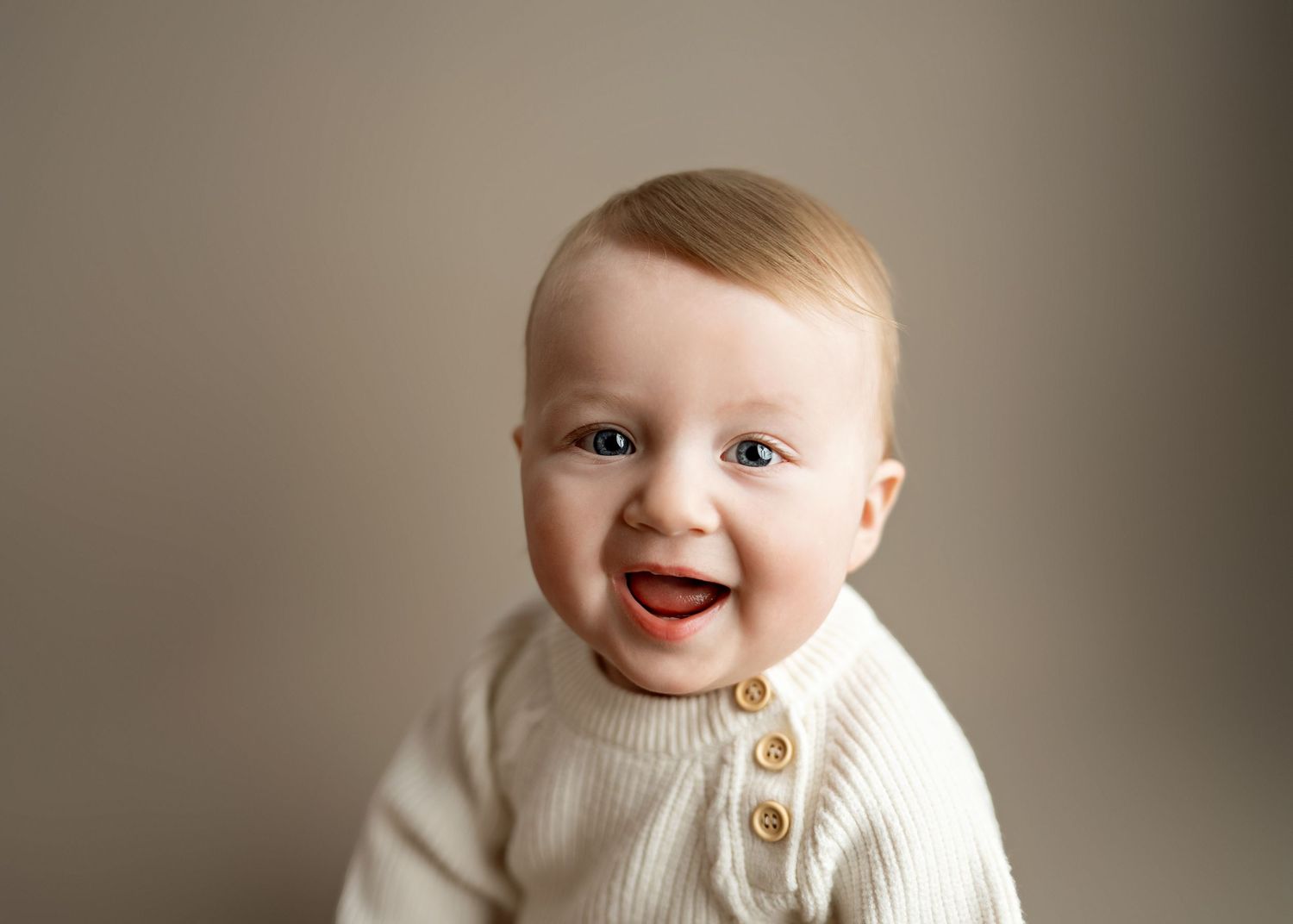 A joyful baby wearing a cream-colored knit cardigan smiles brightly against a neutral background.