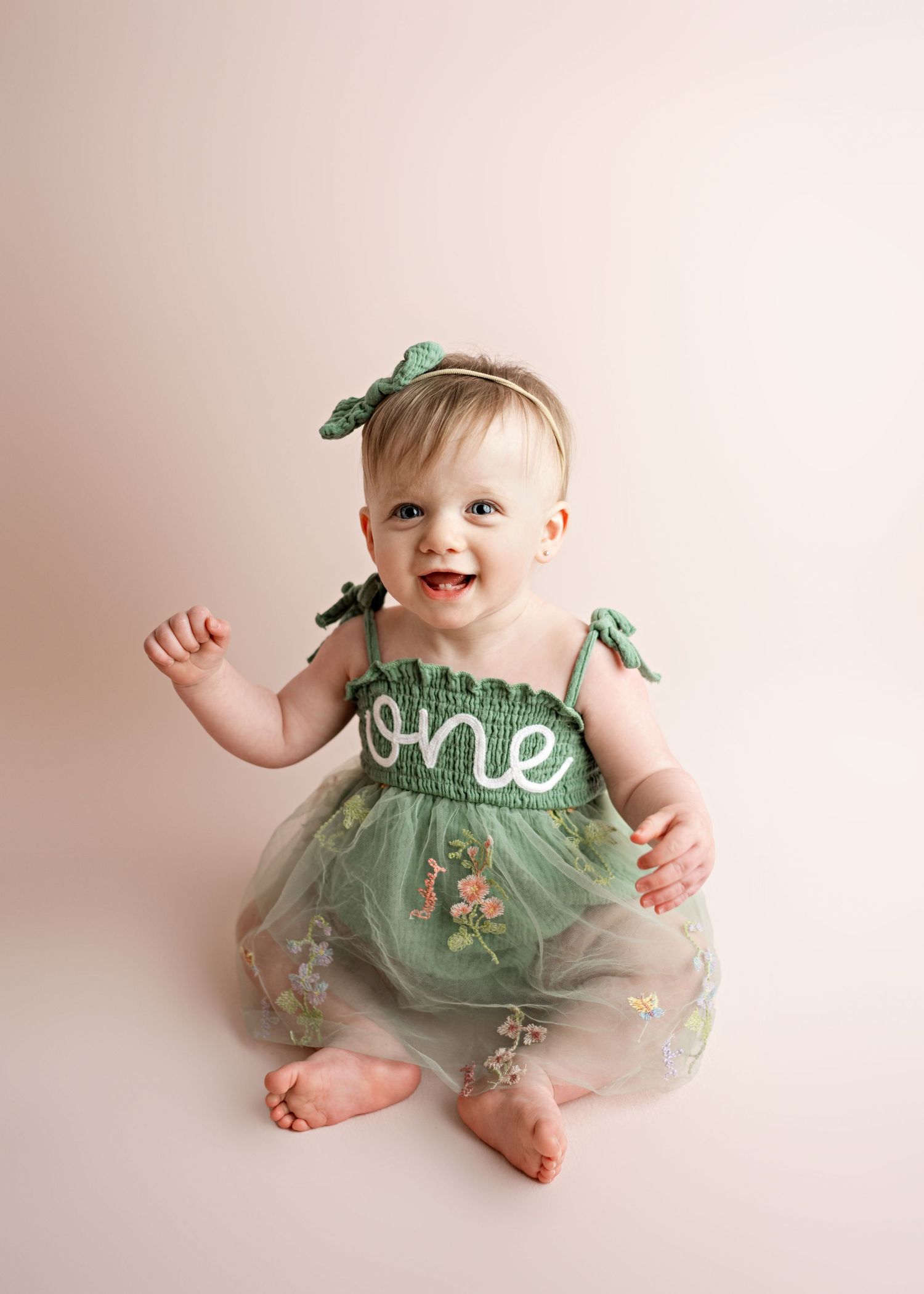 A baby wearing a sage green tulle dress with 'one' text sits smiling against a light pink background.