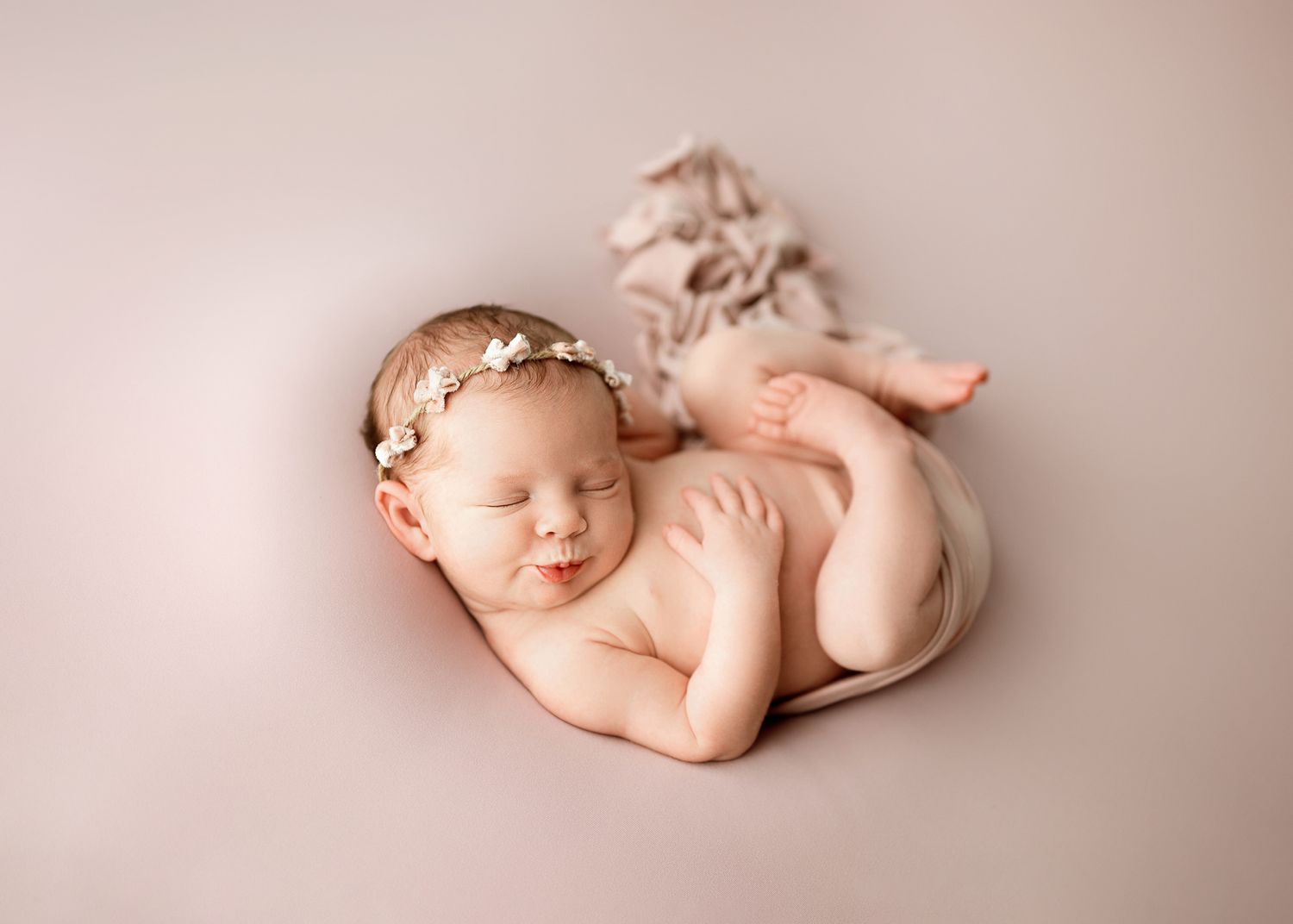 Peaceful sleeping newborn baby wearing delicate floral headband on soft pink backdrop during professional photoshoot.