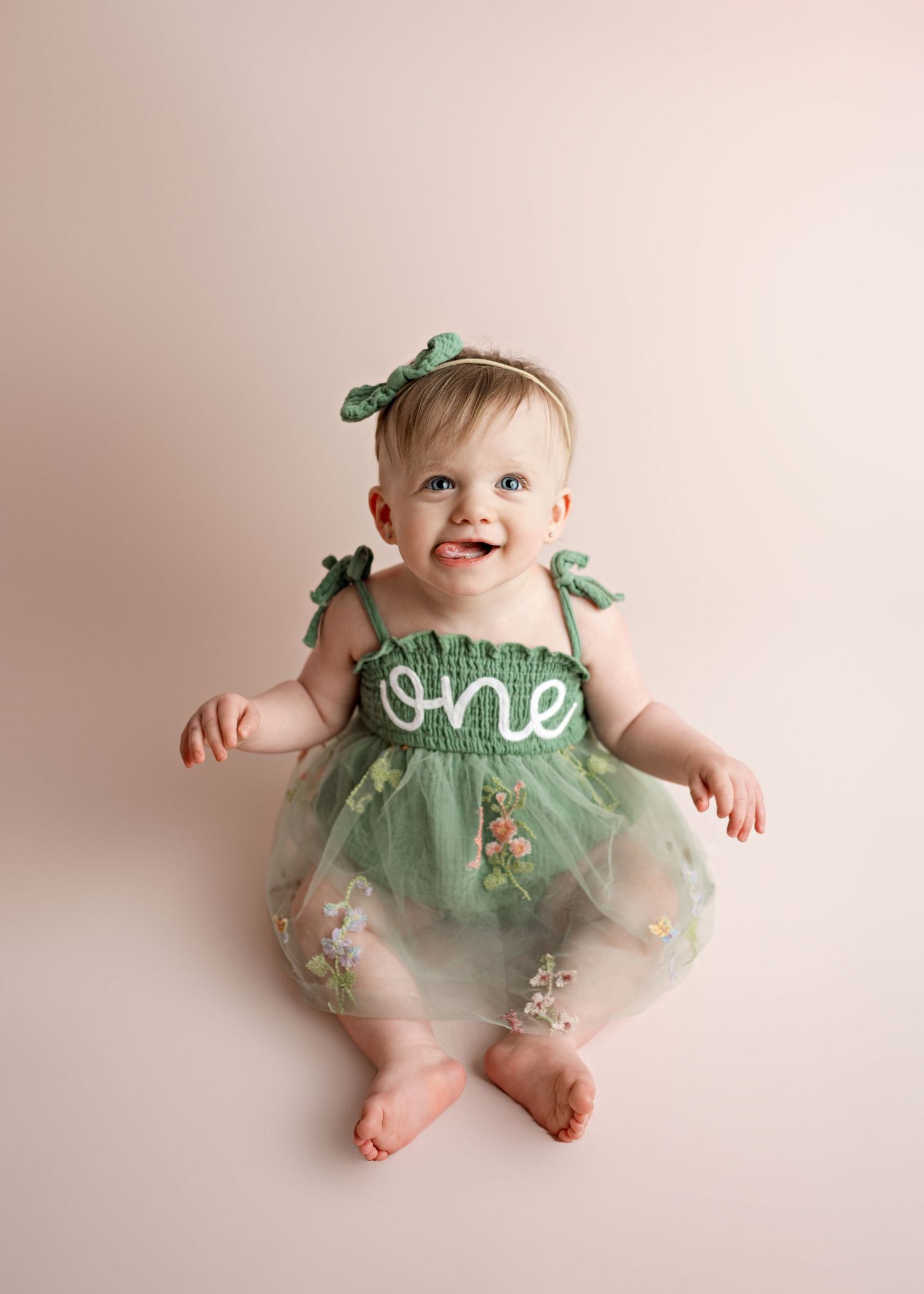 Baby in mint green tulle dress with 'one' text and bows sits on beige background for first birthday photo.