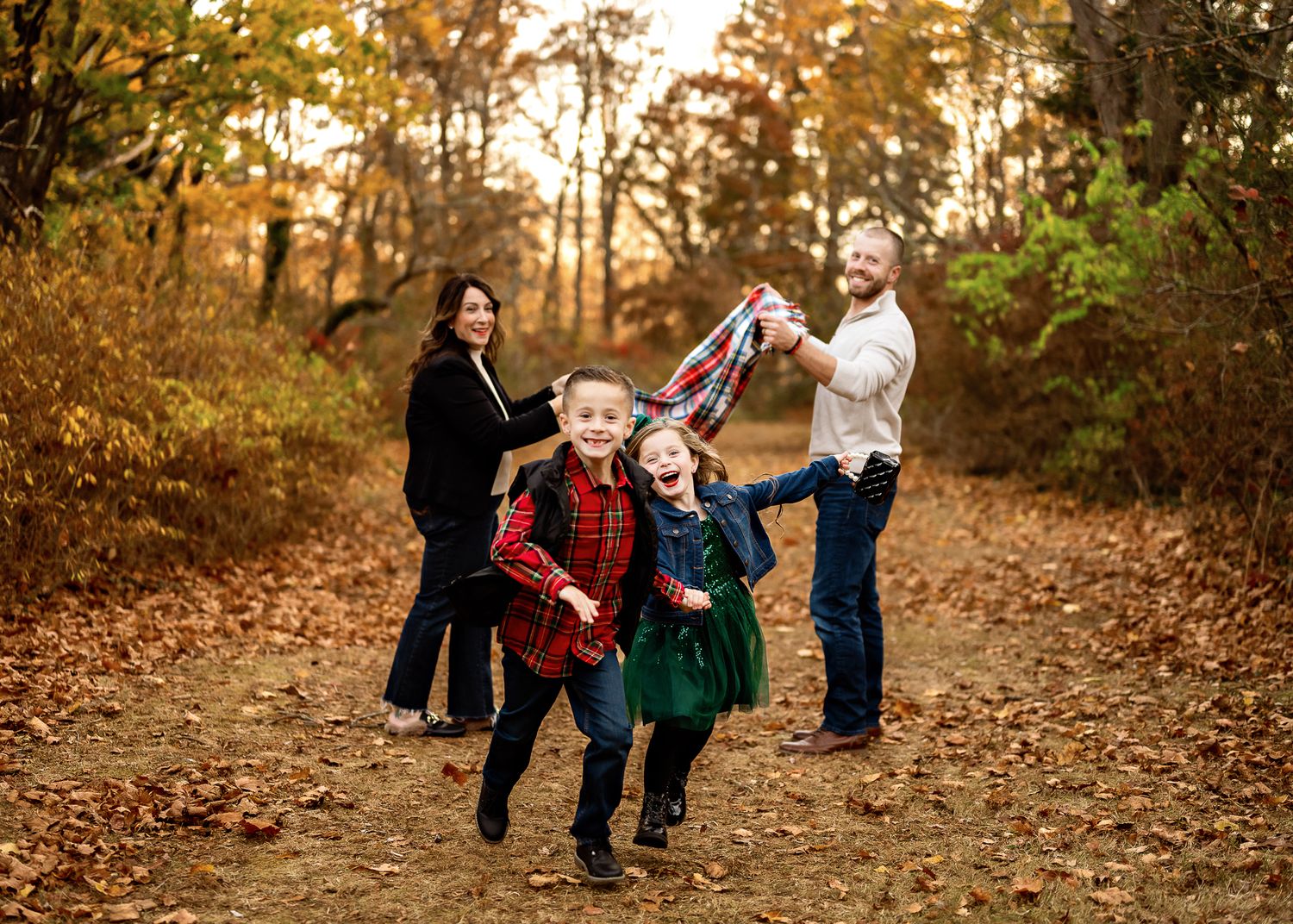 A family enjoys an autumn day playing on a leaf-covered path in the woods during golden hour sunset.