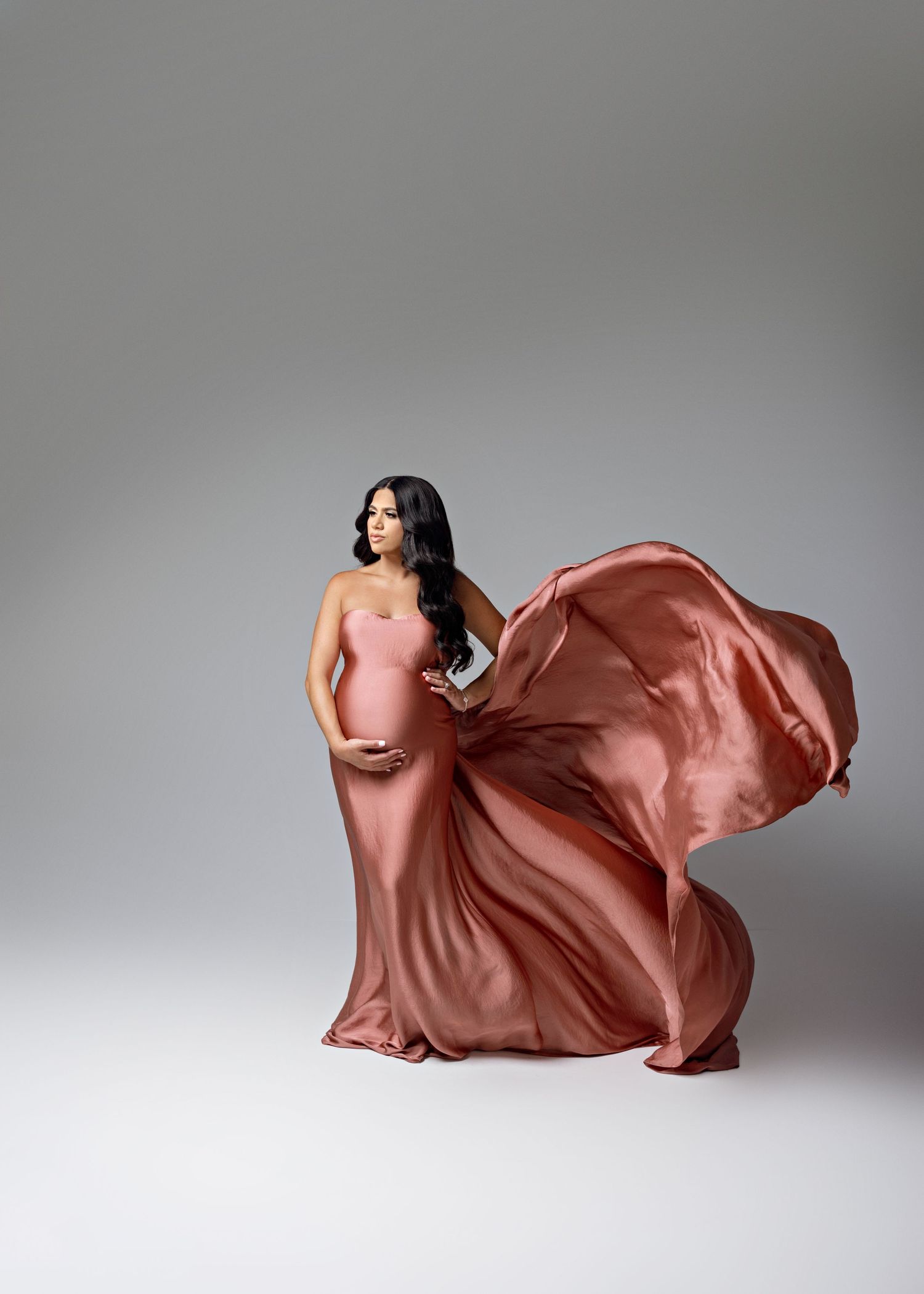 A pregnant woman poses in a flowing rose gold silk gown against a gray backdrop as the fabric billows dramatically.