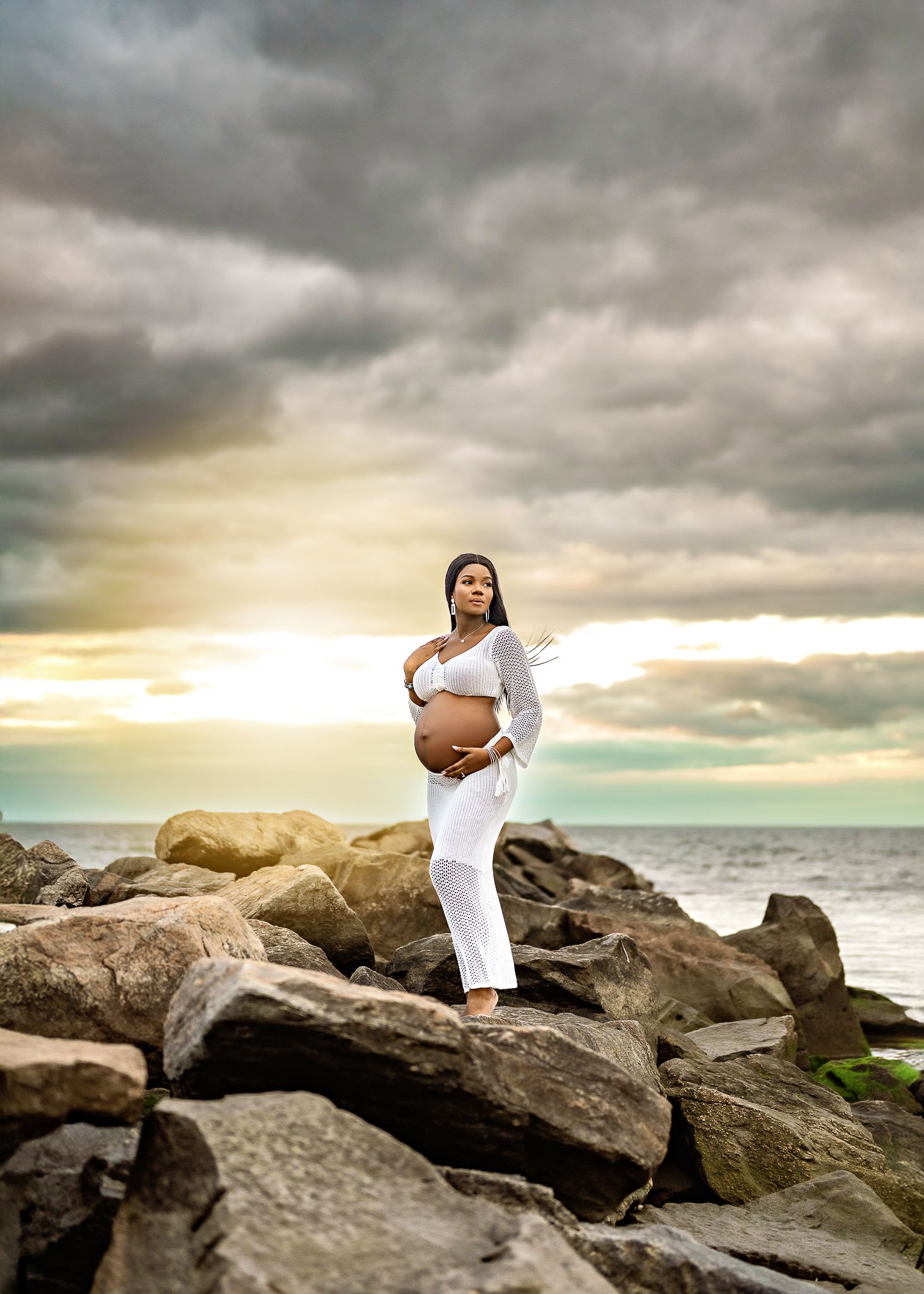A pregnant woman in white clothing poses gracefully on coastal rocks at sunset with dramatic cloudy skies overhead.