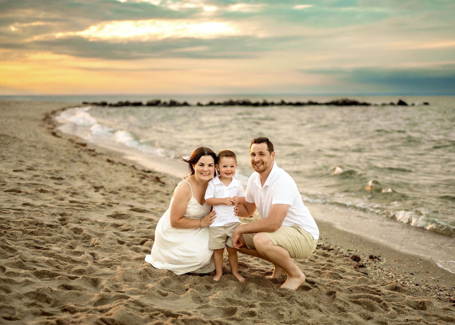 Three individuals in white attire sit together on a sandy beach at sunset with ocean waves in the background.