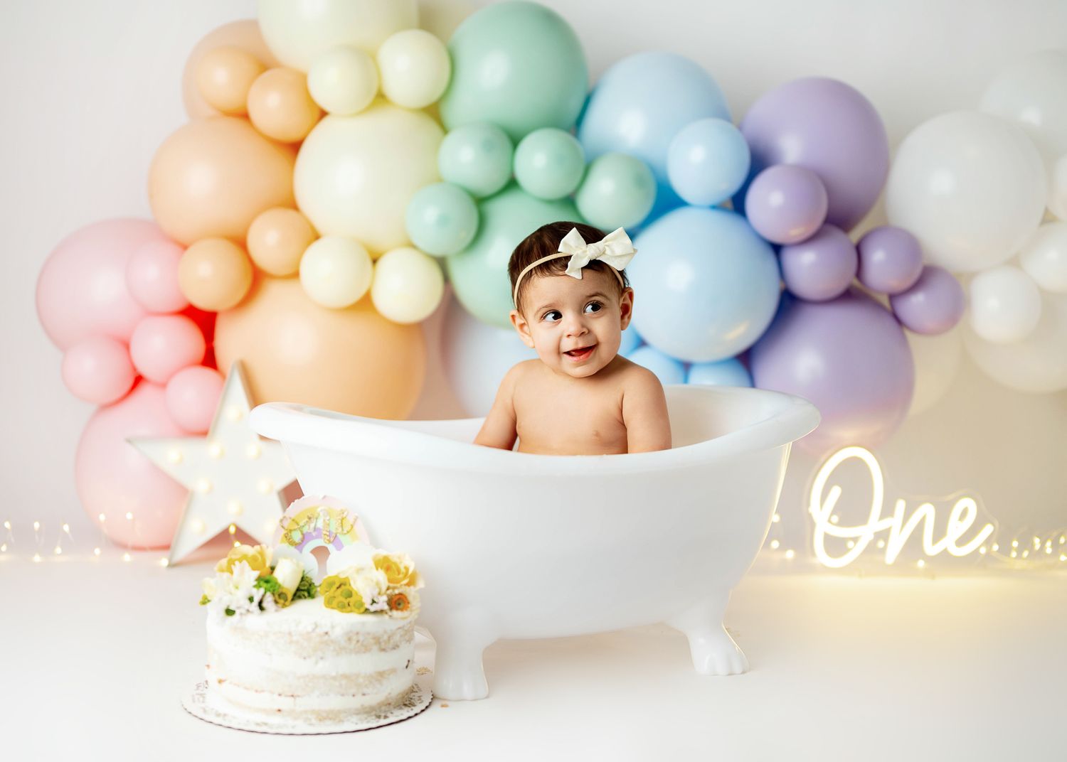 Pastel balloon arch decorations in pink, blue, mint and yellow colors frame a birthday cake setup against a light background.