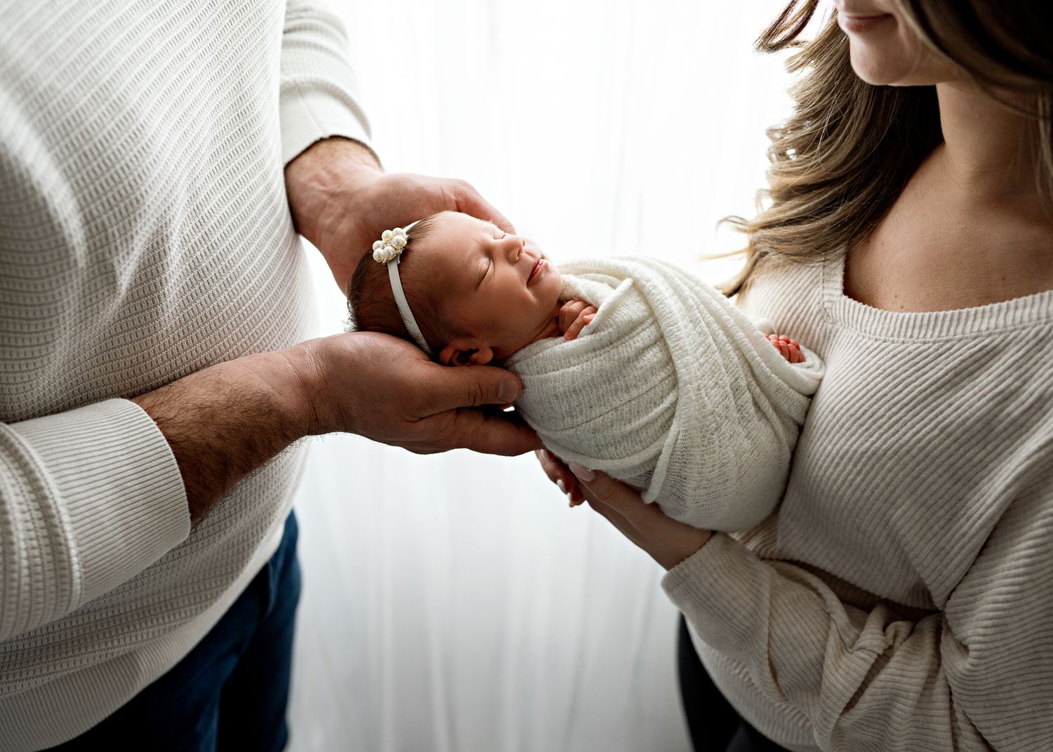 A sleeping newborn swaddled in a light-colored blanket is cradled gently between two people wearing neutral knits.