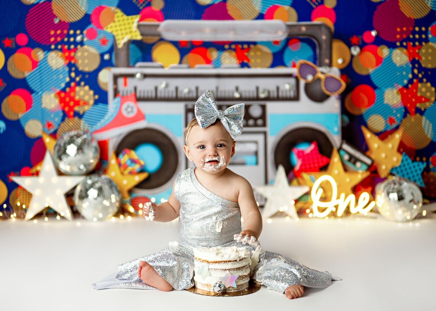 Baby in silver sparkly outfit sits with cake against colorful disco party backdrop with glowing boombox decorations.