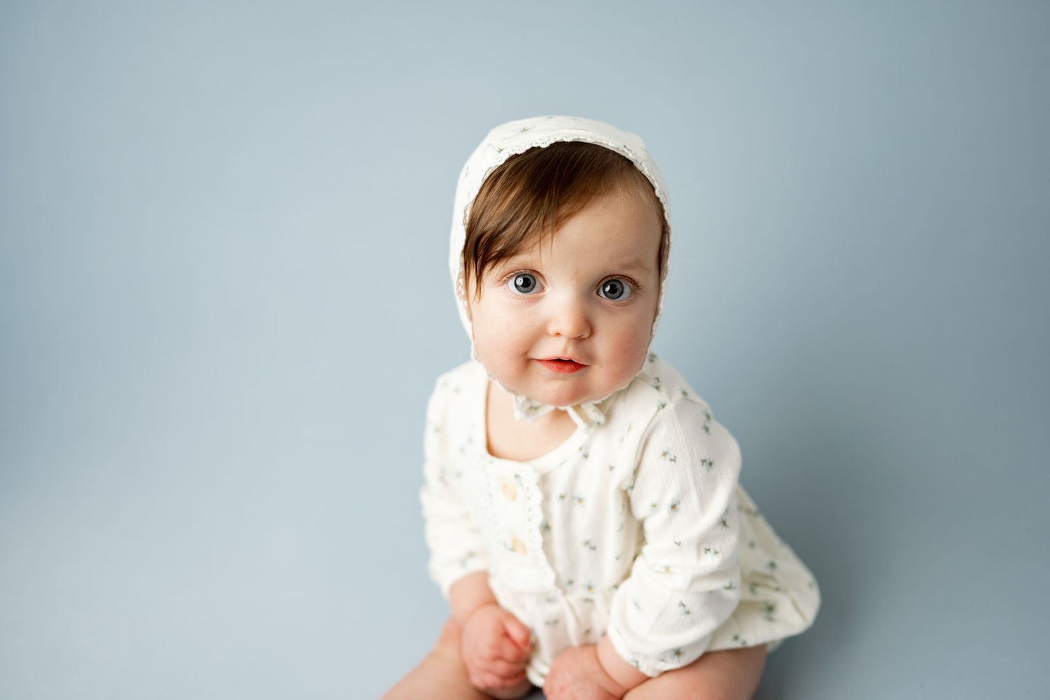 A baby in a white knit sweater sits against a light gray background looking at the camera.