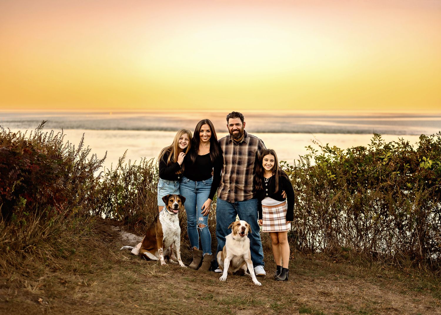 Group poses with two dogs on coastal bluff during golden sunset at beach.