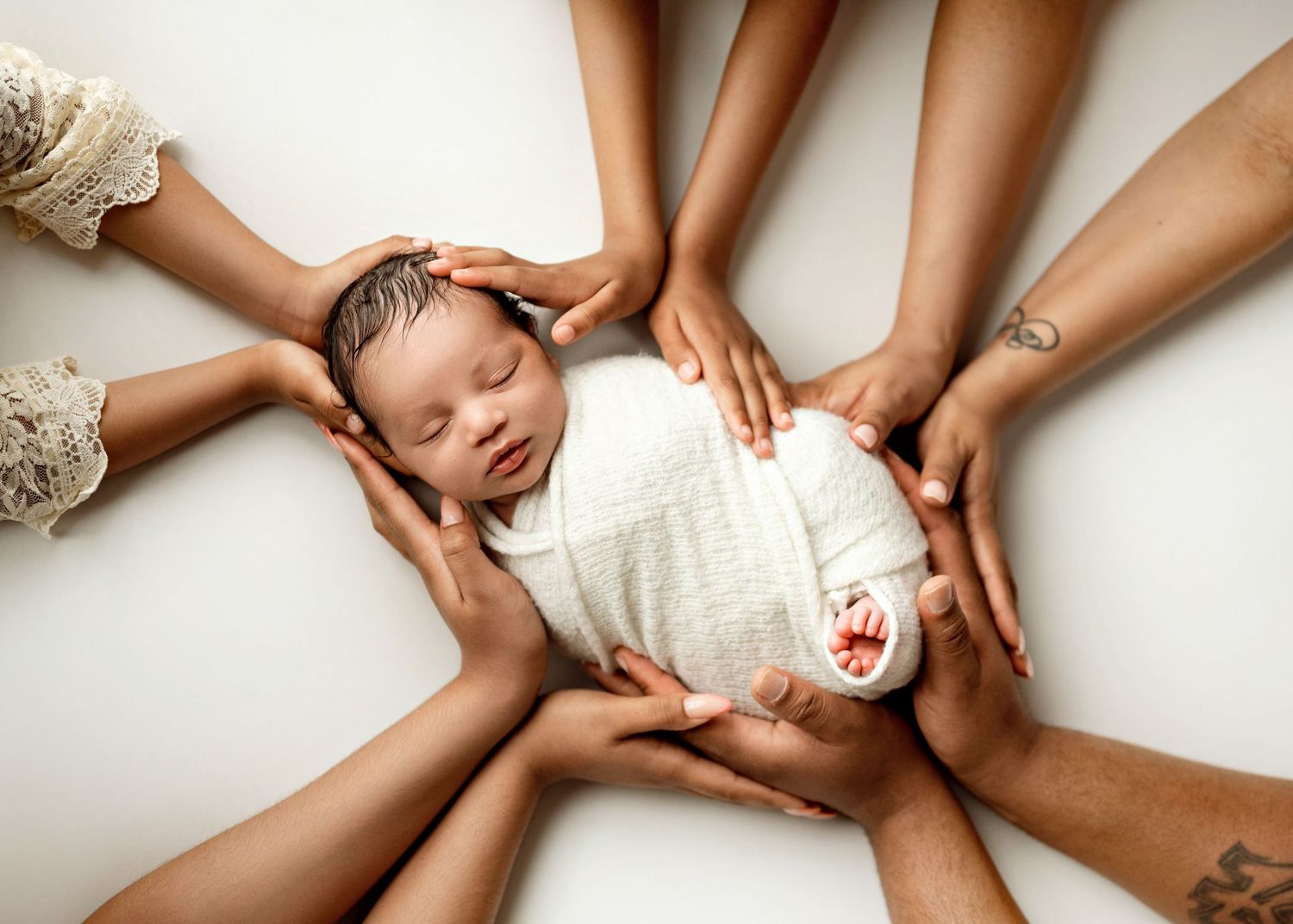 Multiple hands surround a sleeping newborn wrapped in white swaddling cloth against a light background.