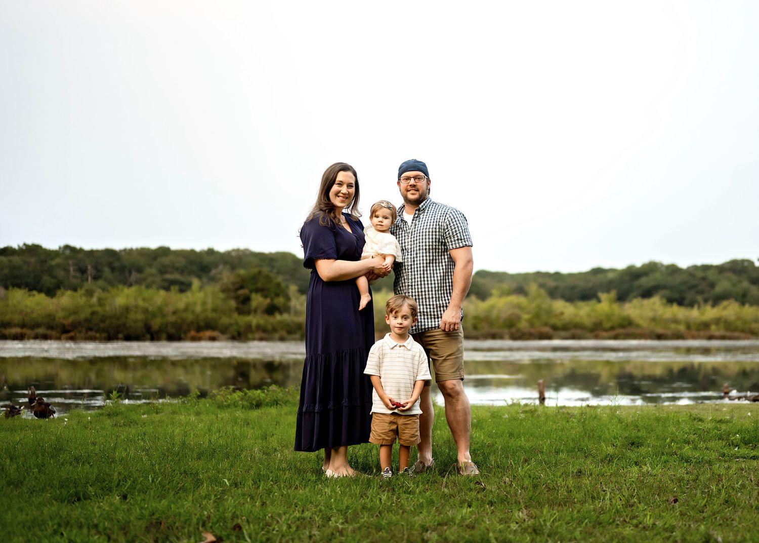 A family stands together on green grass with a marsh and trees visible in the background.