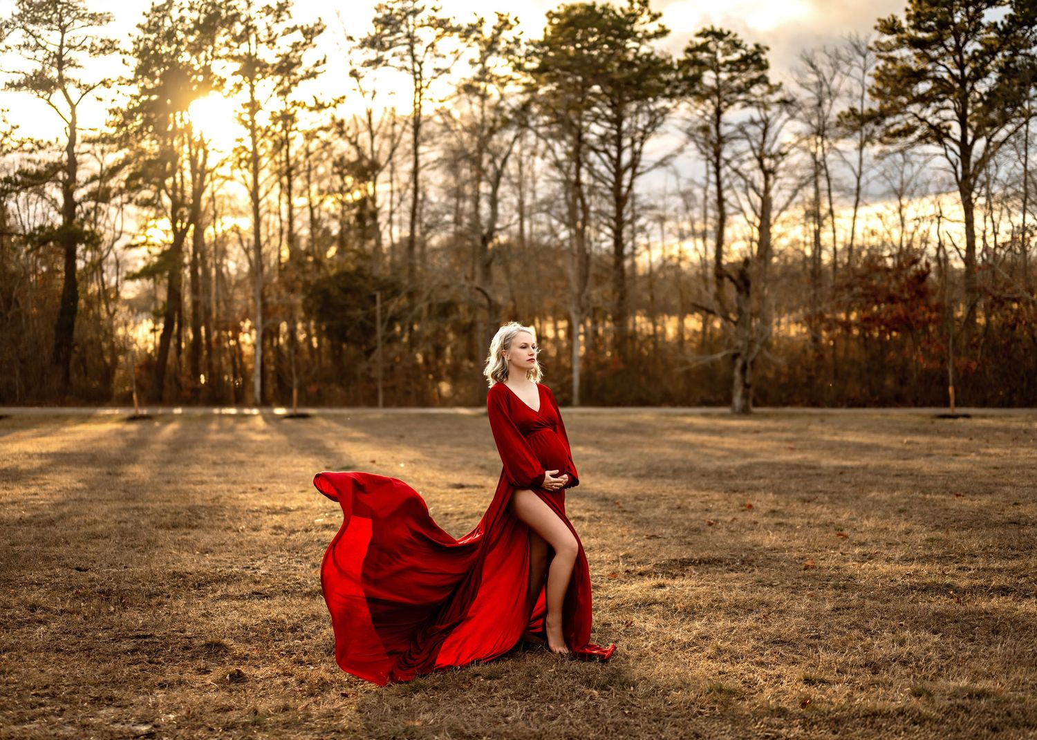 A flowing red dress billows dramatically in a field at sunset with sunlight streaming through tall pine trees.