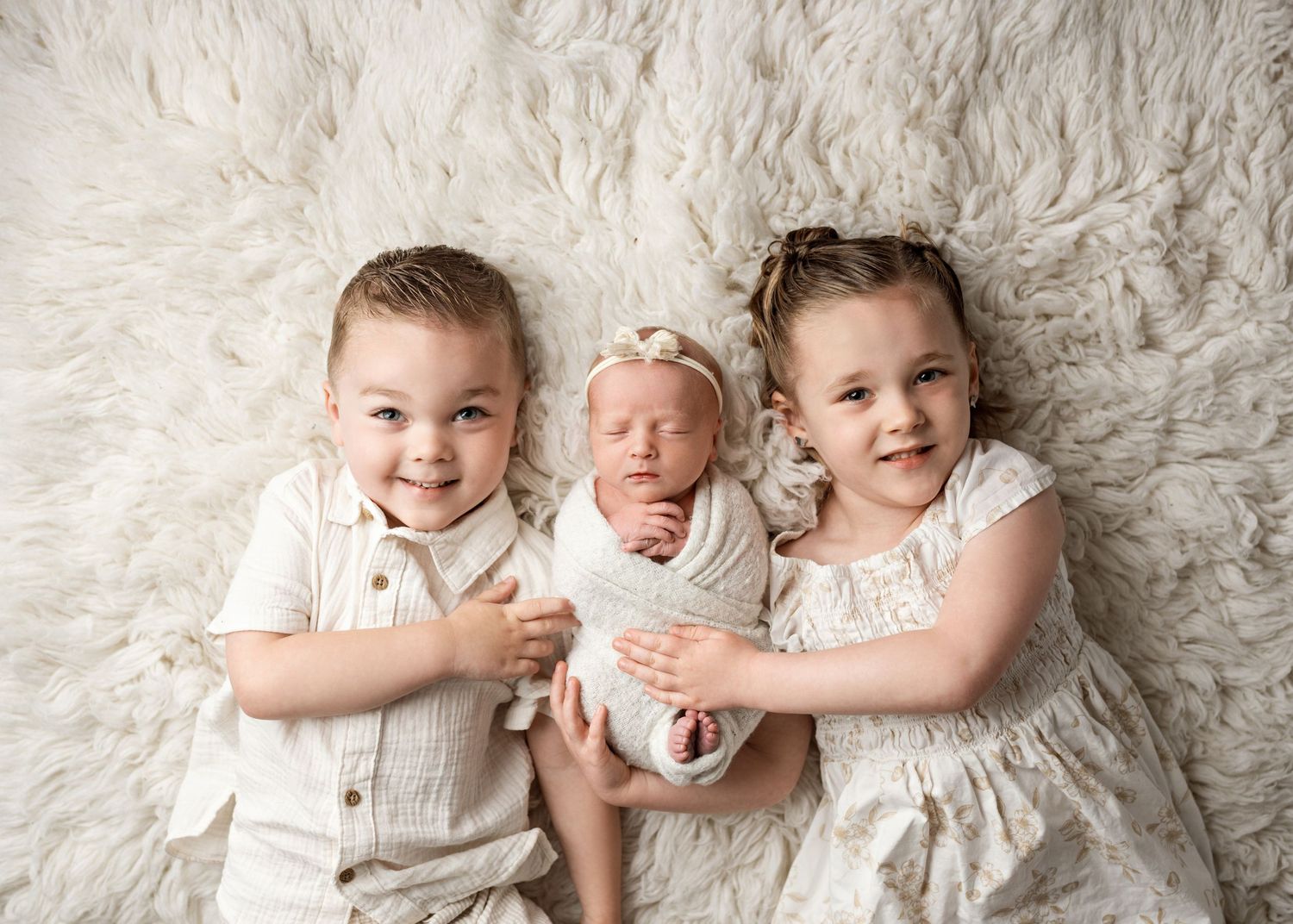 Three siblings in white outfits lying together on a cream-colored furry rug for a portrait session.