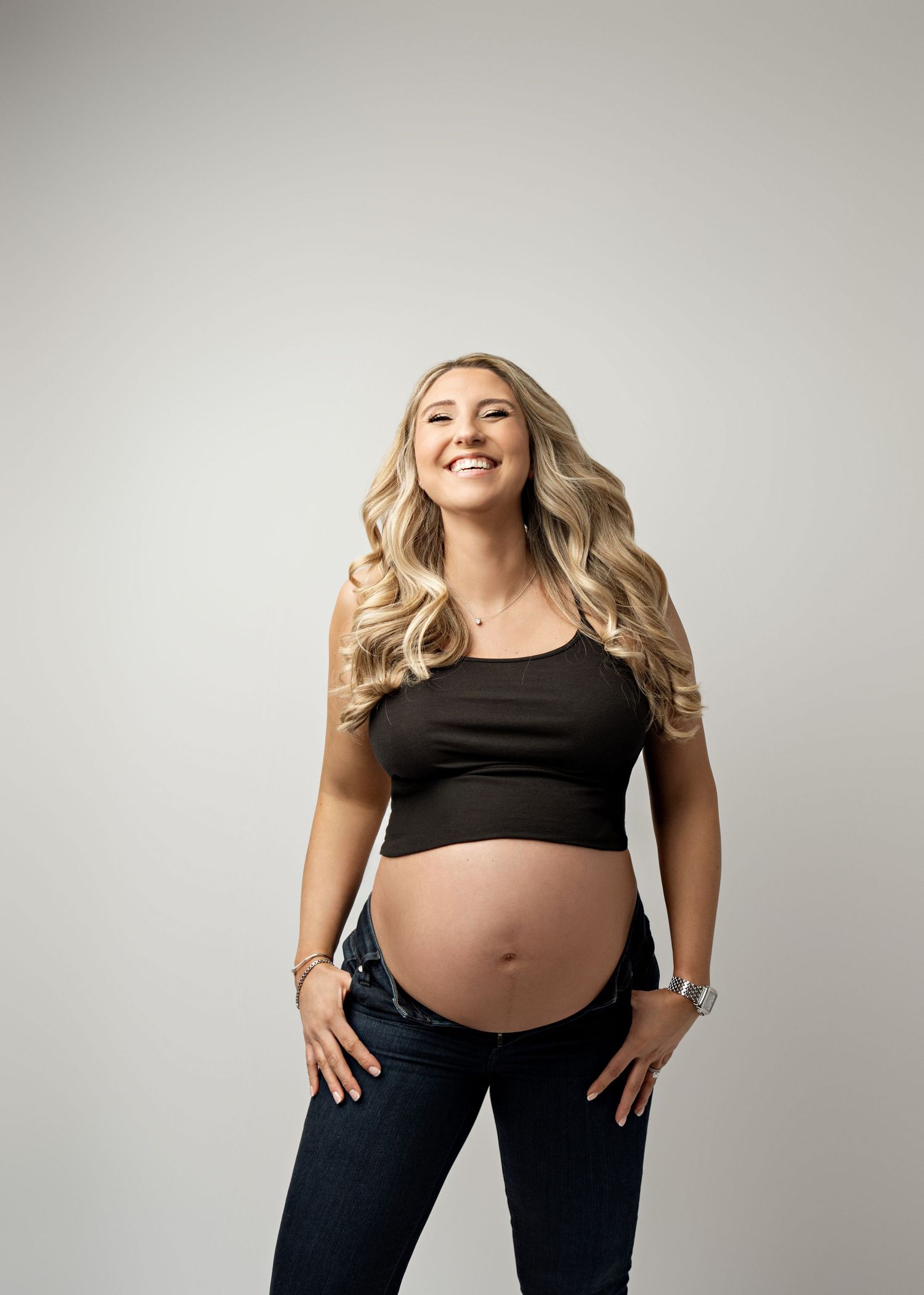 A pregnant woman in a black crop top and jeans poses against a neutral background with a bright smile.