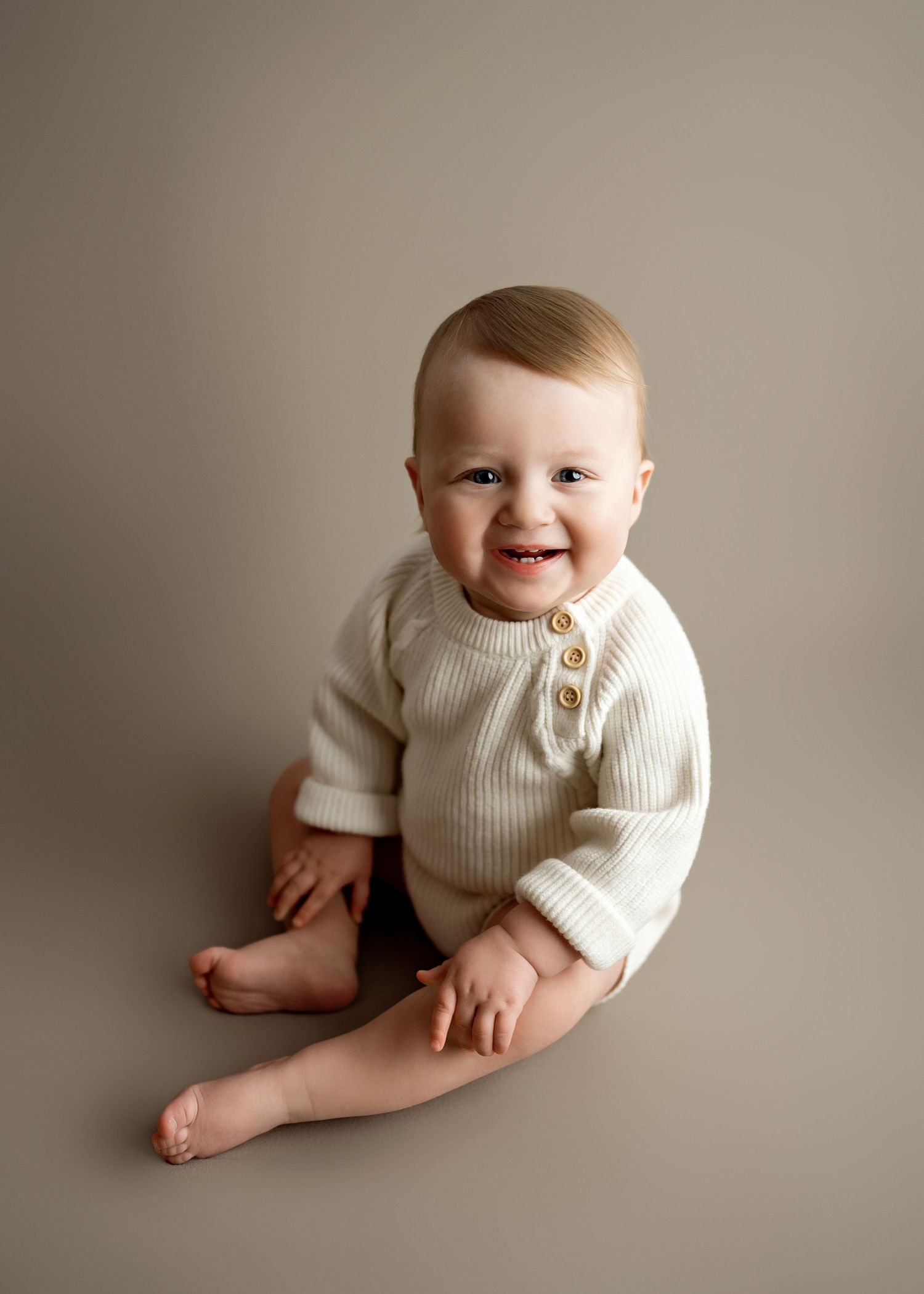 Smiling baby wearing cream ribbed sweater sits against neutral backdrop in minimalist portrait style.