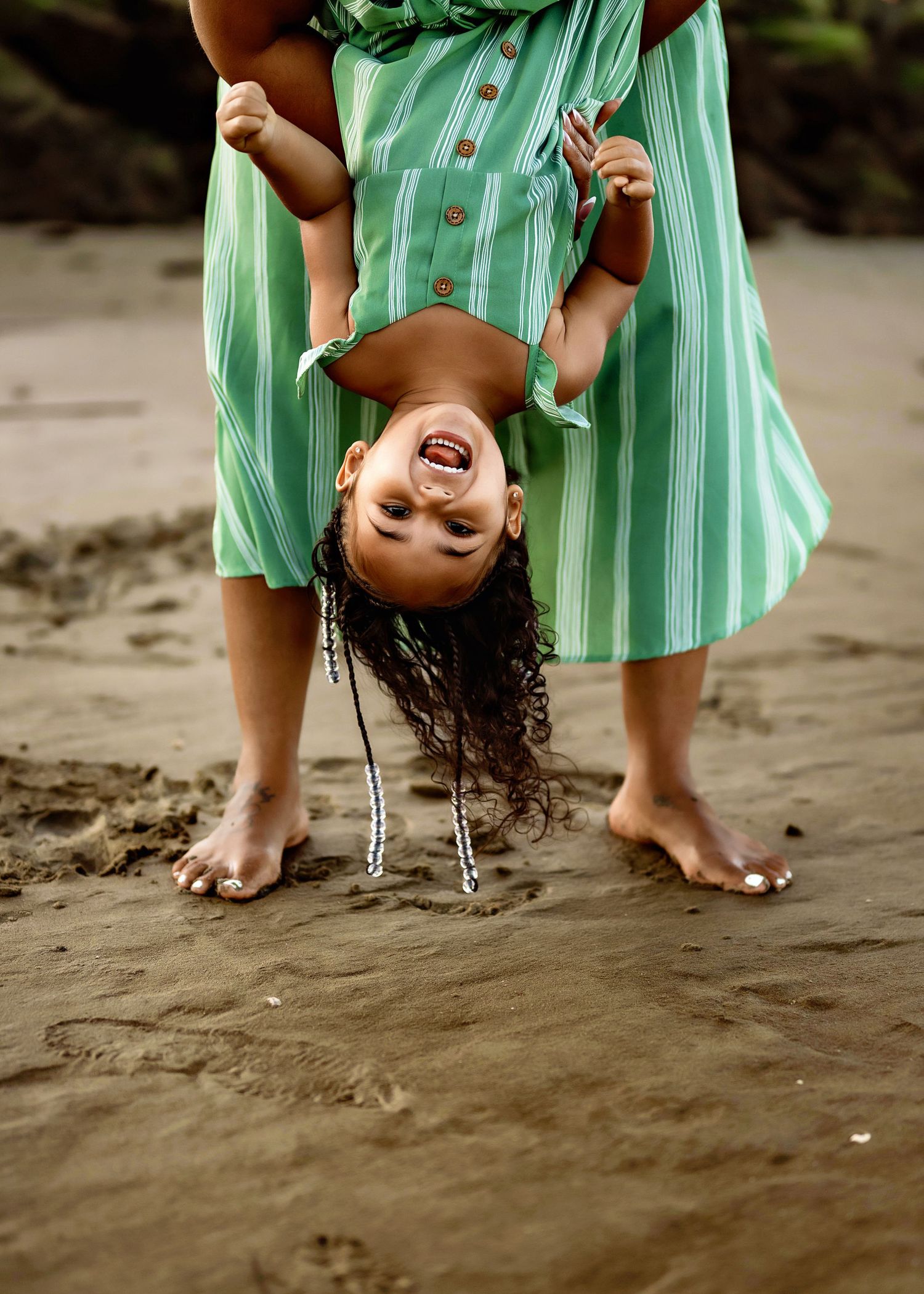 Person wearing mint green striped dress holds child upside down on sandy beach, both laughing and enjoying playful moment.