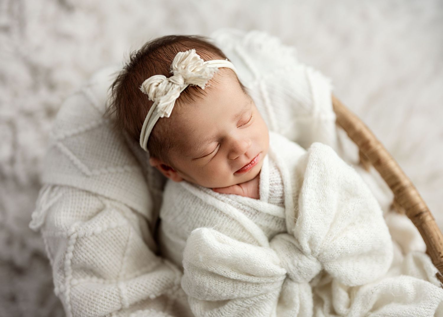Sleeping newborn in soft white swaddle blanket and lace headband rests peacefully in wooden basket.