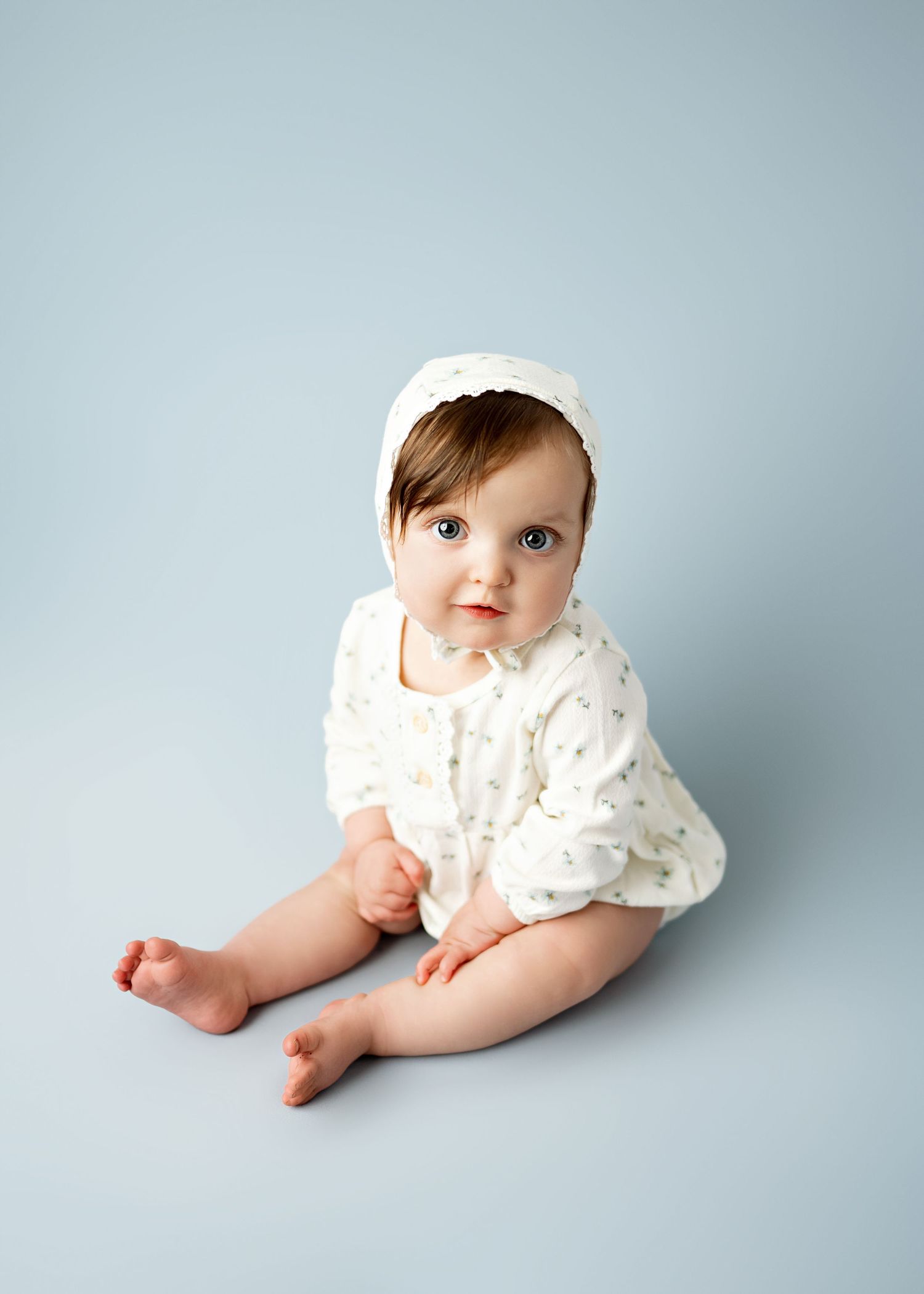 Baby wearing white bonnet and floral romper sits on light gray background looking at camera.