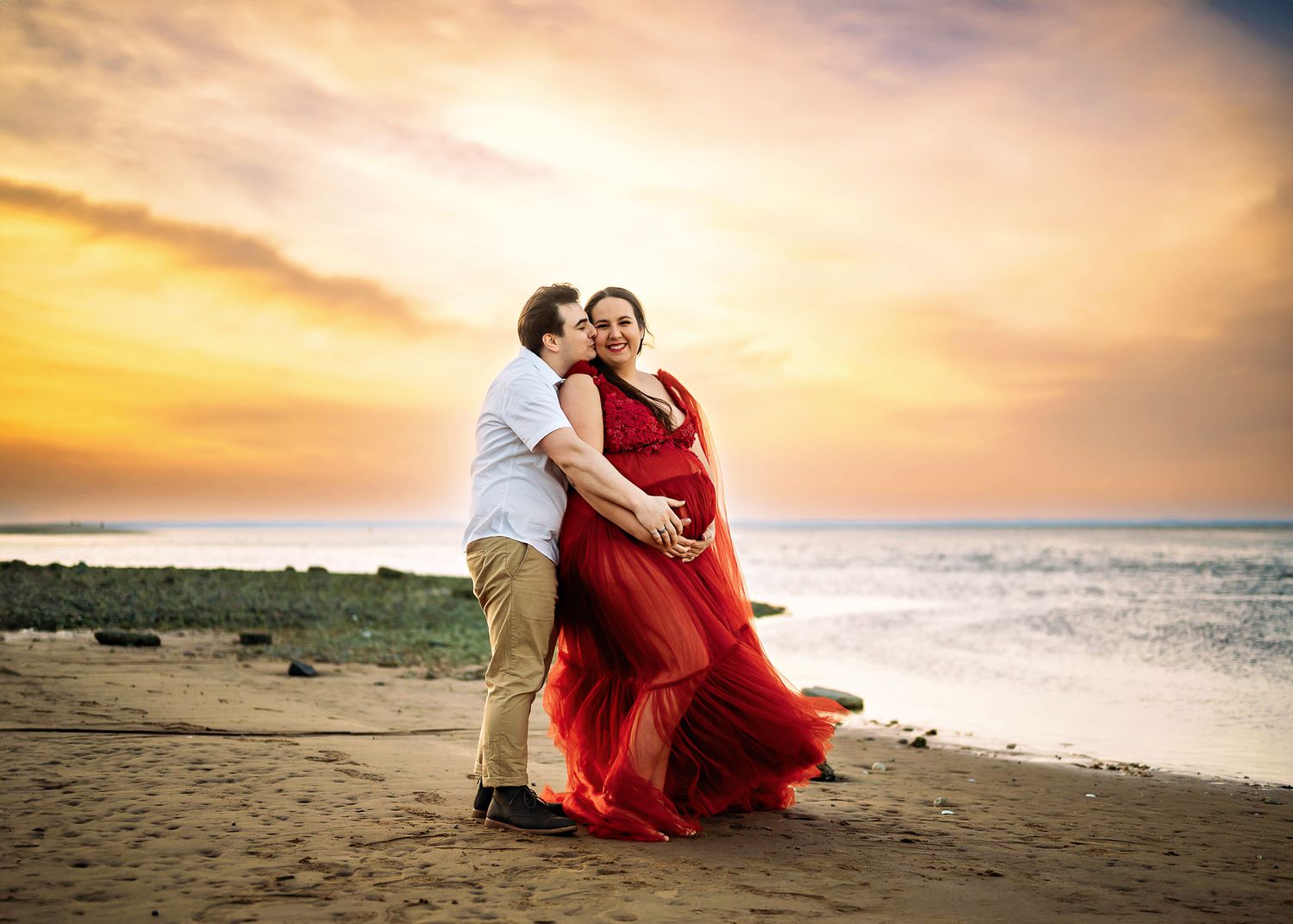 A romantic couple poses on the beach at sunset, with the woman wearing a flowing red dress against the golden sky.