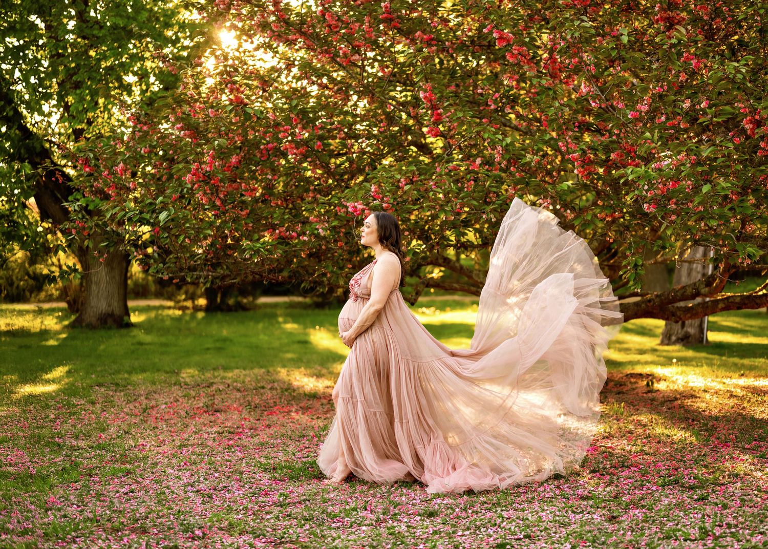 A flowing pink gown billows in a spring garden with blooming cherry blossoms and golden sunlight filtering through trees.