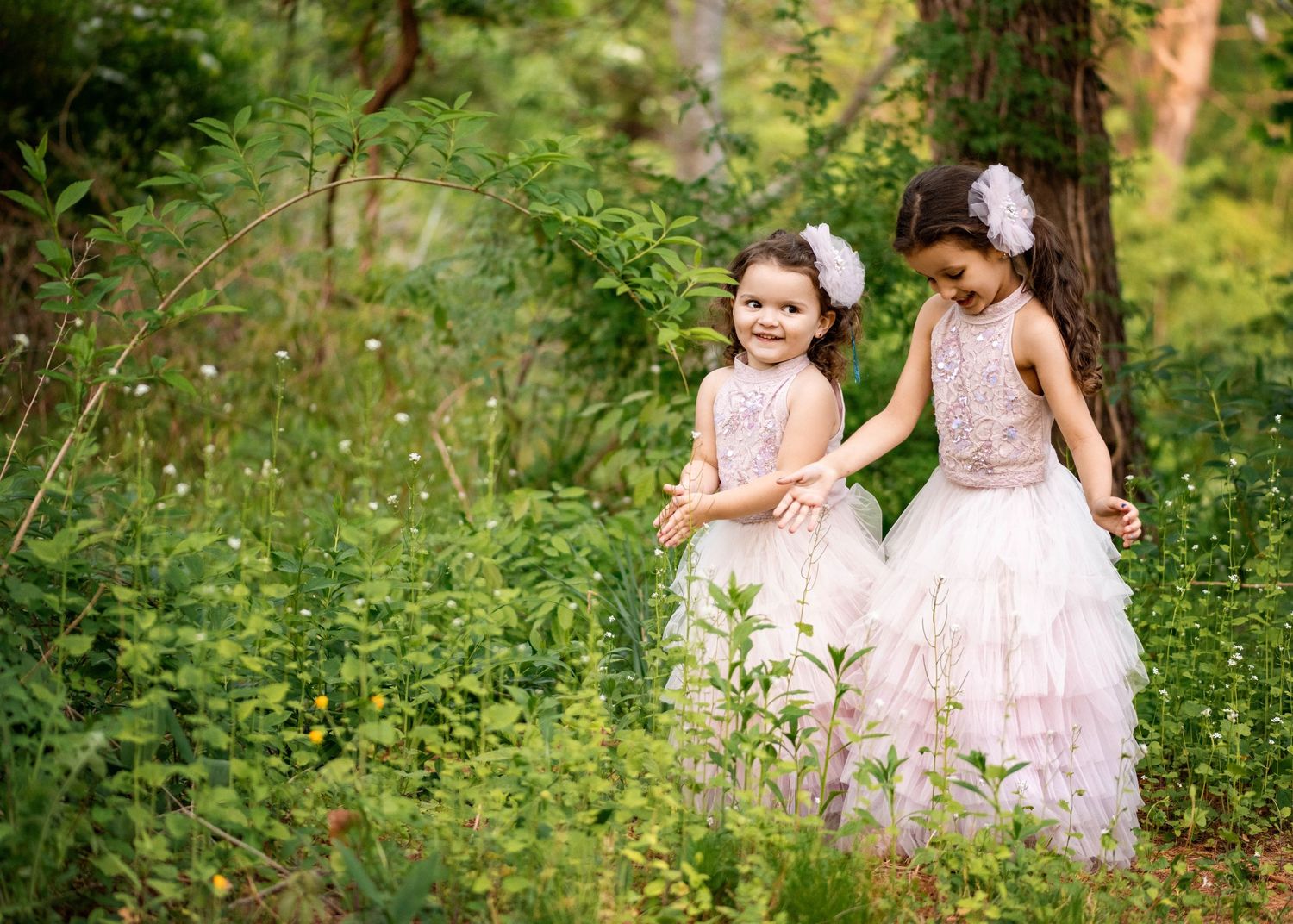 Two young girls in pink dresses walk together through a lush green forest setting in summer.