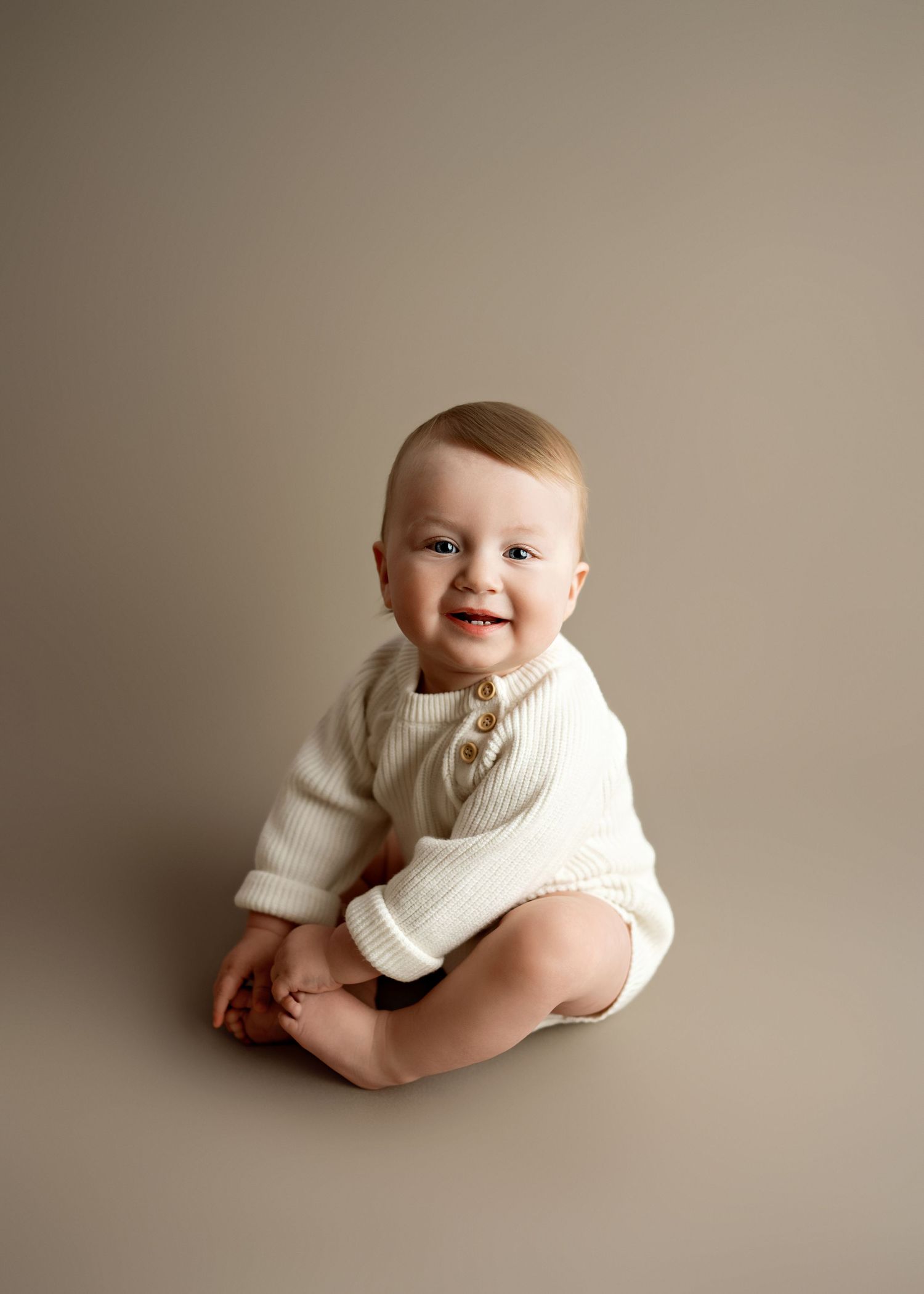 A smiling baby wearing a cream knit sweater sits on a neutral-toned backdrop.