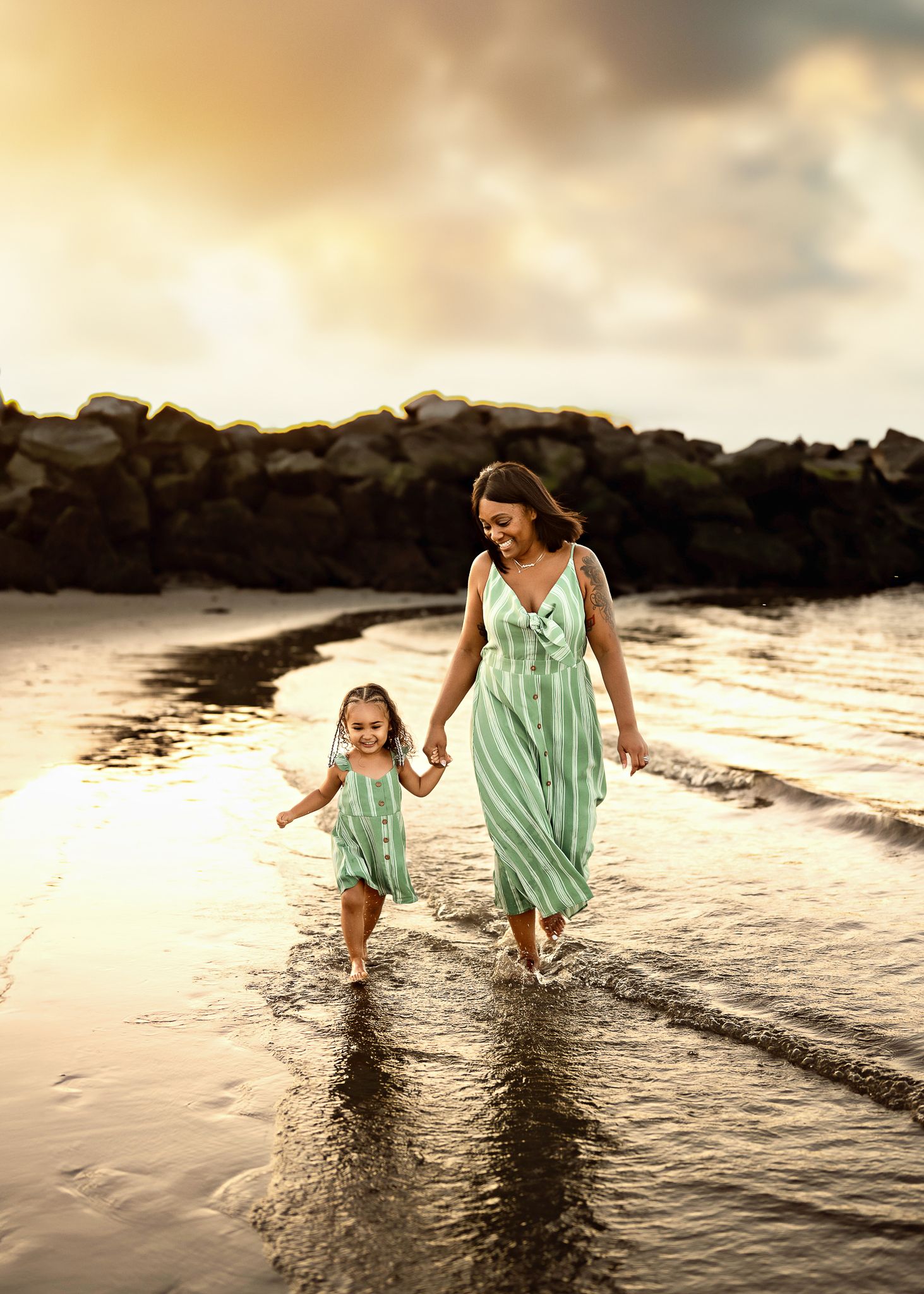 Two people in matching mint green dresses walk hand in hand along a sunset beach with rocks in the background.