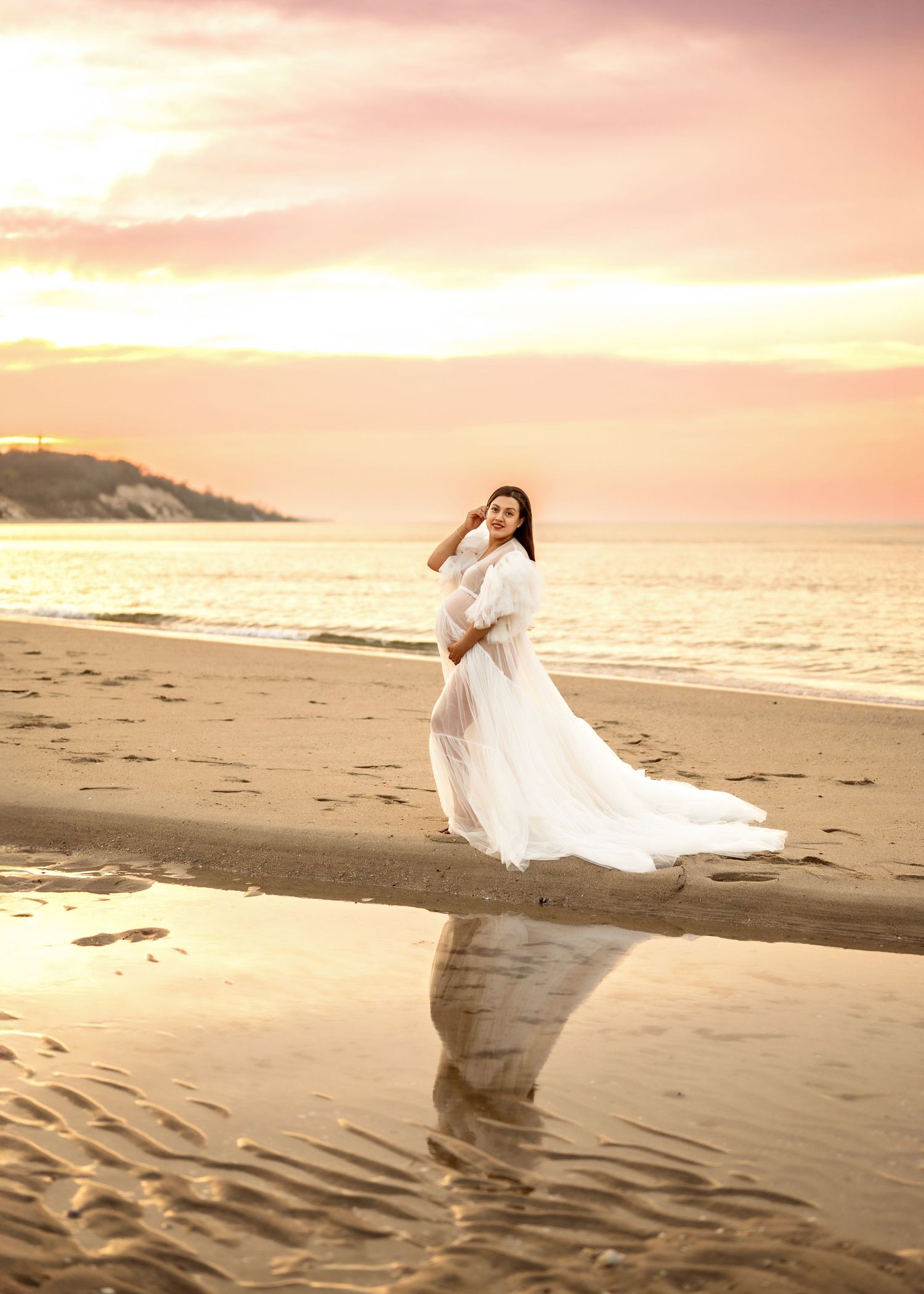 A person in a flowing white dress poses on a beach during golden sunset with waves gently lapping at the shore.