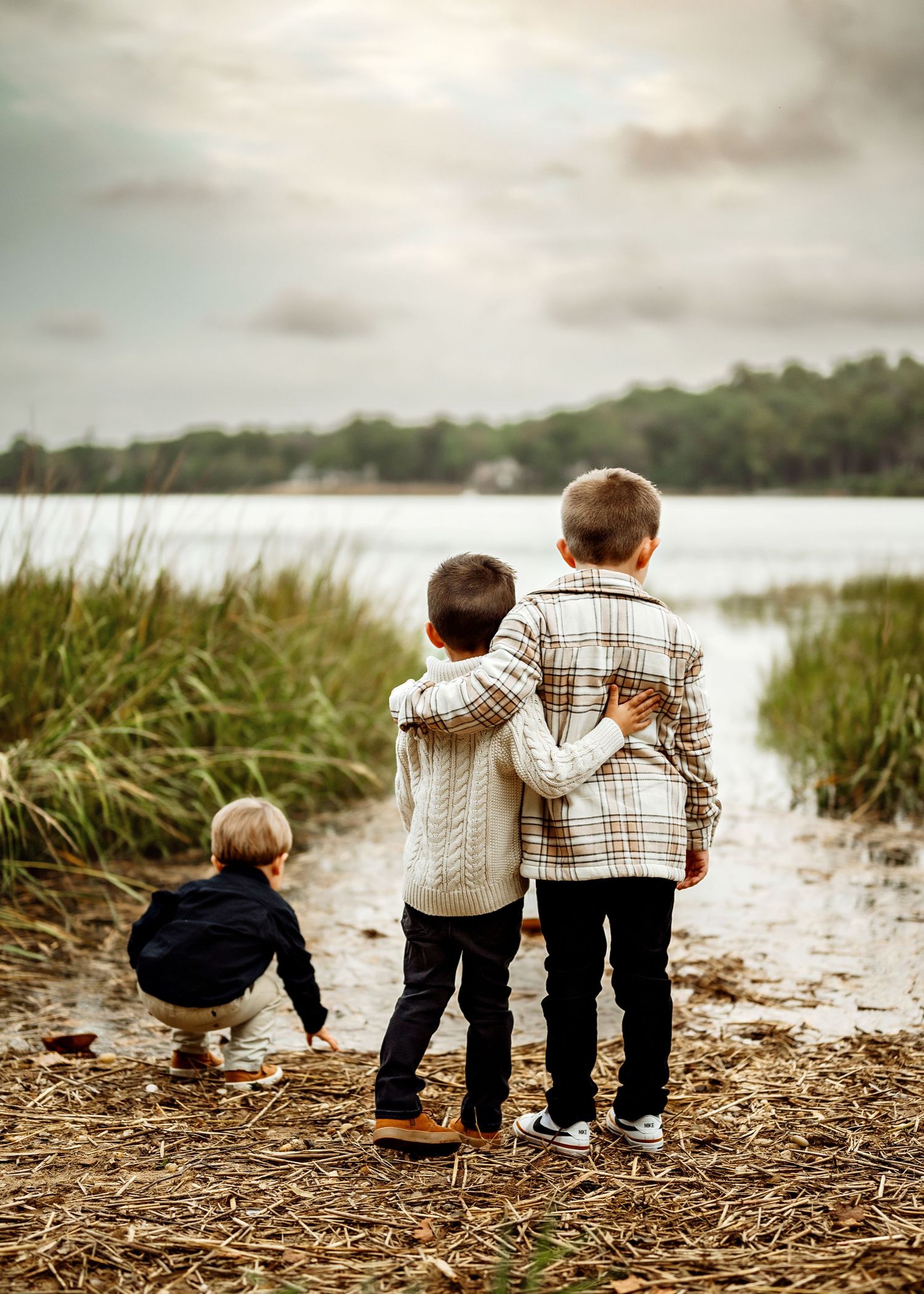 Three children stand together on a lakeside path surrounded by tall grass while looking out at the peaceful water.