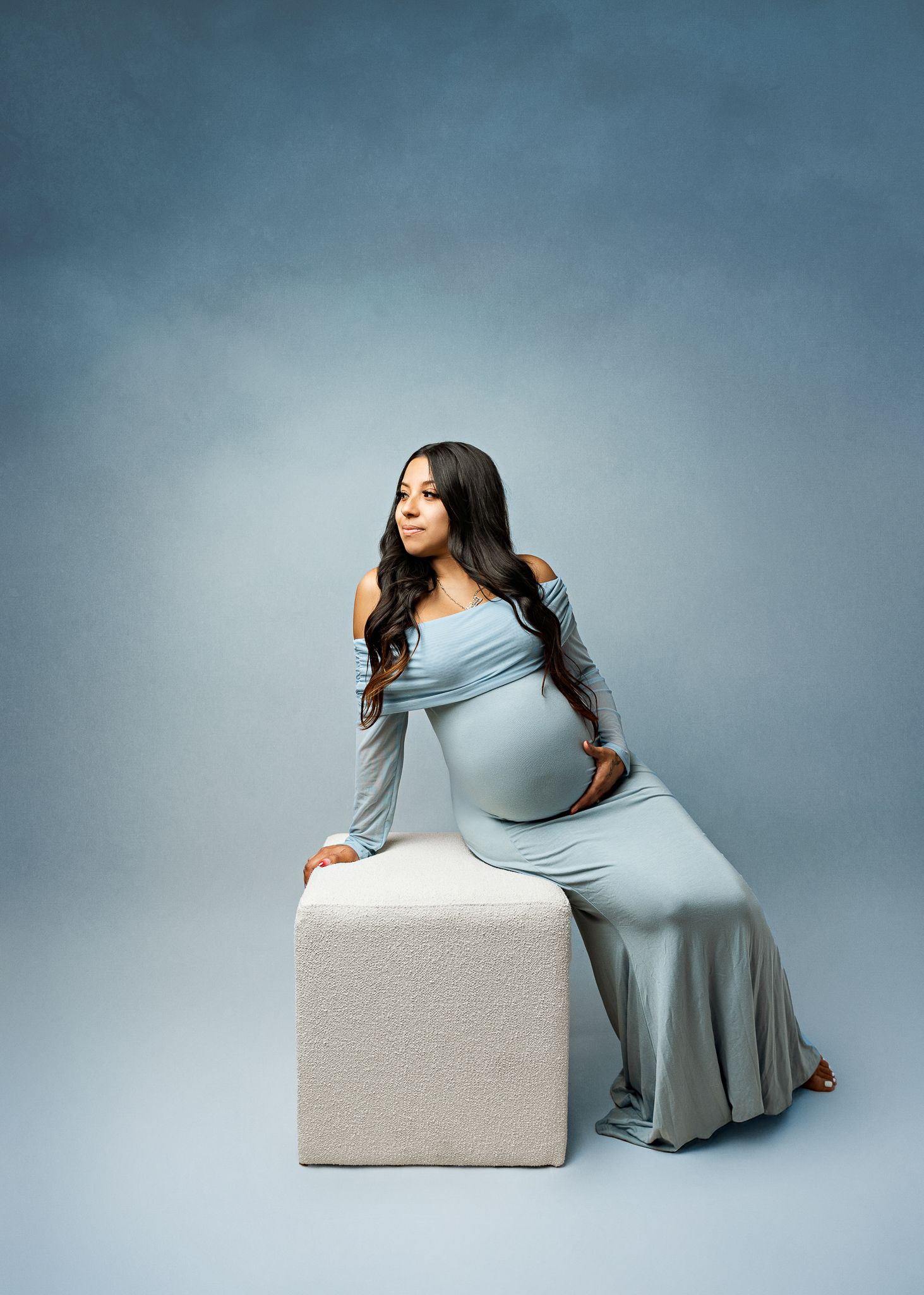 A pregnant person in a flowing blue dress poses elegantly on a white cube against a gray studio backdrop.