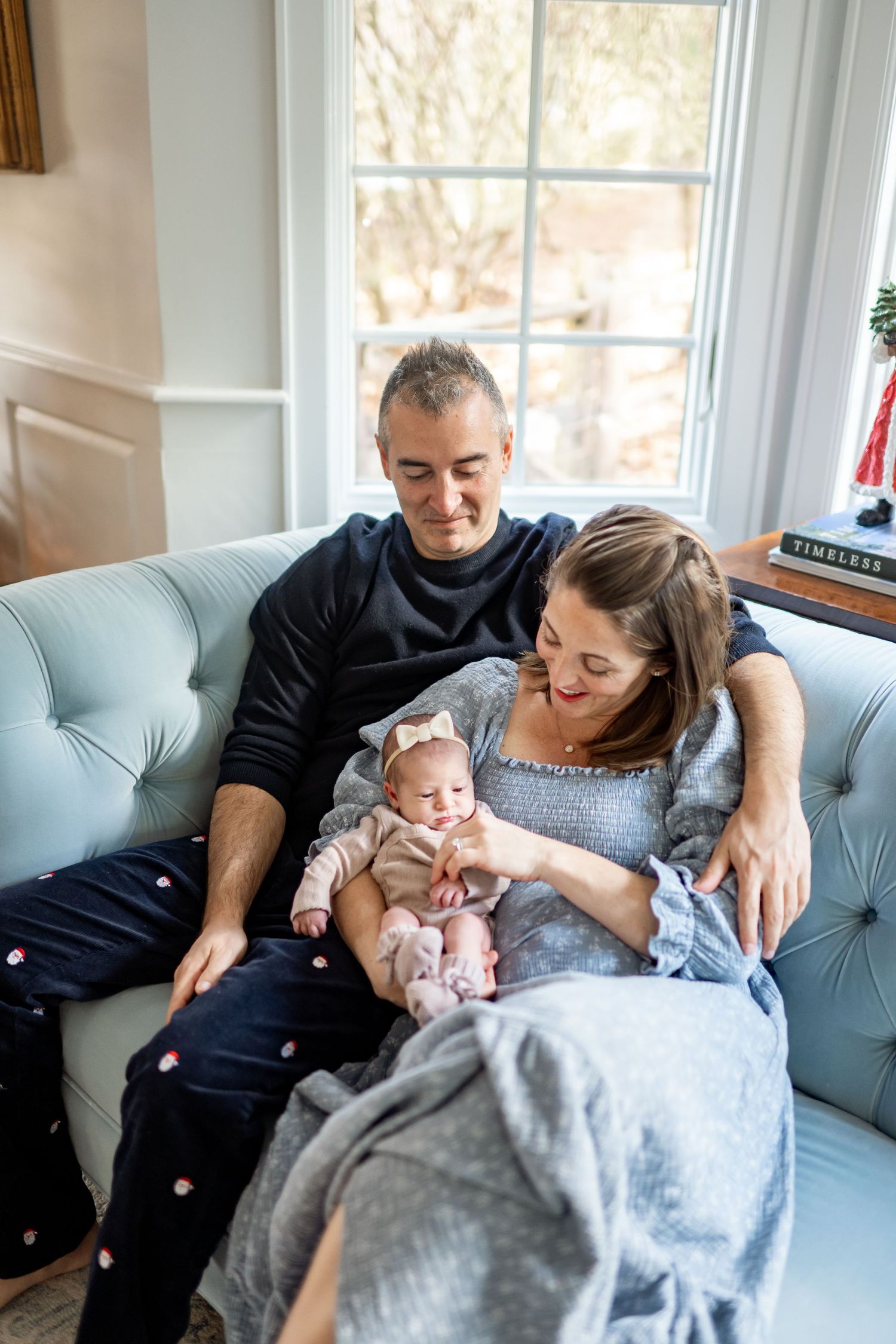 A couple sitting on a light blue couch lovingly gazes at their infant baby at home near a bright window.