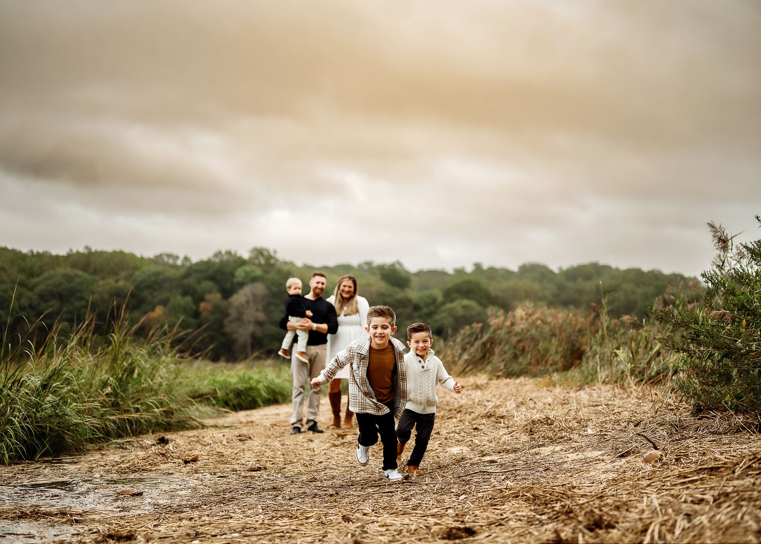 A group of children run down a dirt path through tall grass at sunset in warm golden light.