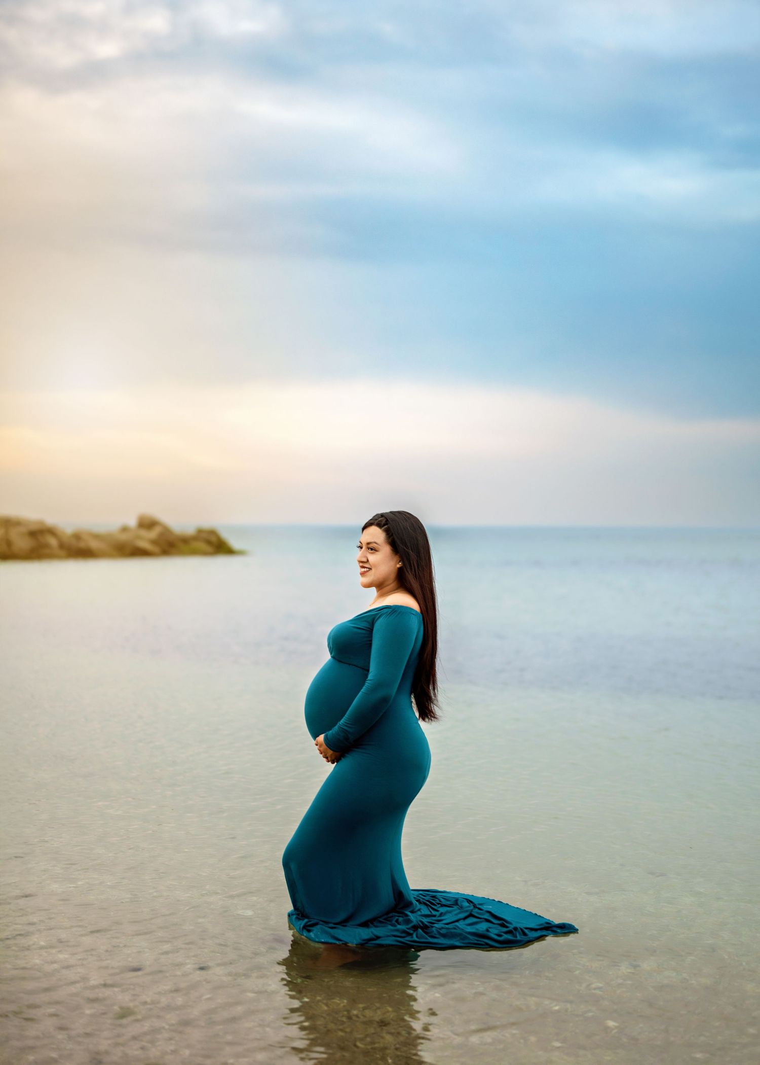 A person in a teal maternity gown stands in shallow water at sunset against a serene ocean backdrop.