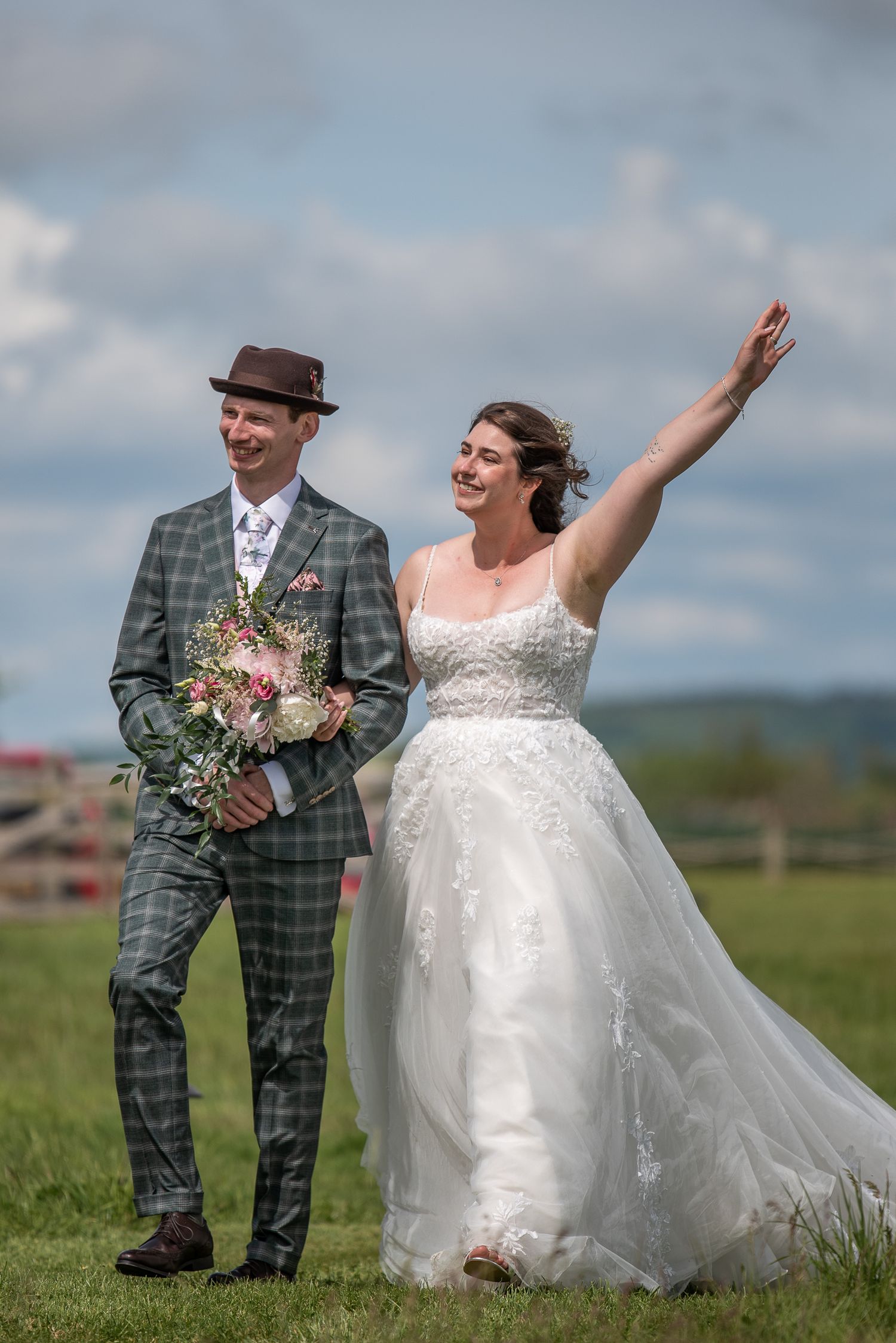 Bride and groom walk in to greet their guests in a field at reception. It's sunny, they're smiling and the bride has one hand in the air