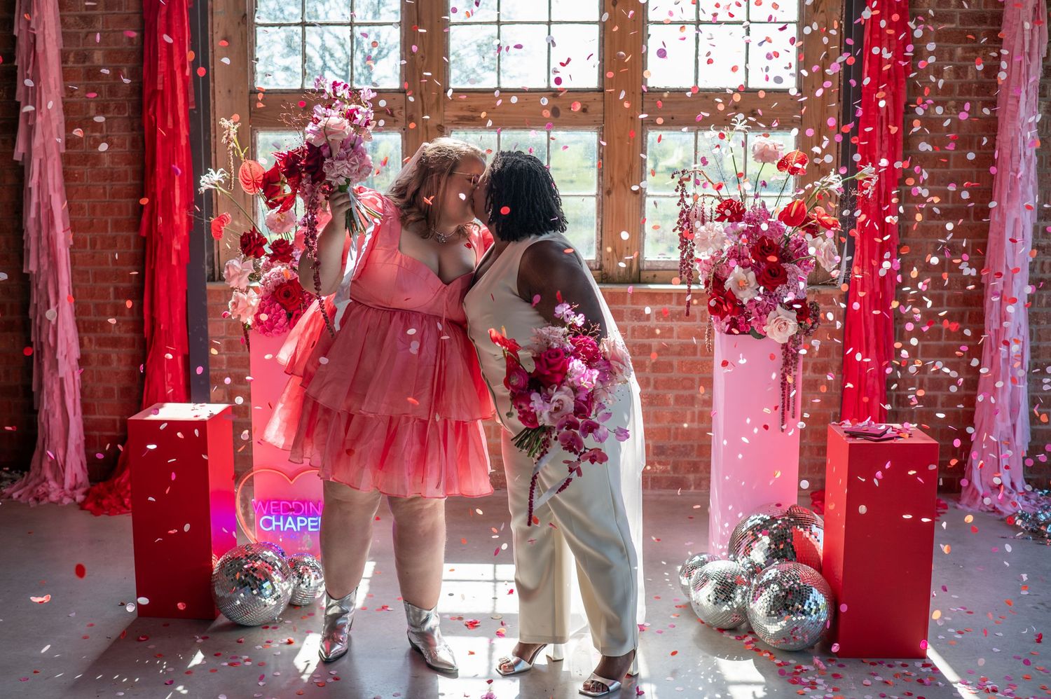 Two brides kiss as beautiful pink and peach paper confetti falls around them