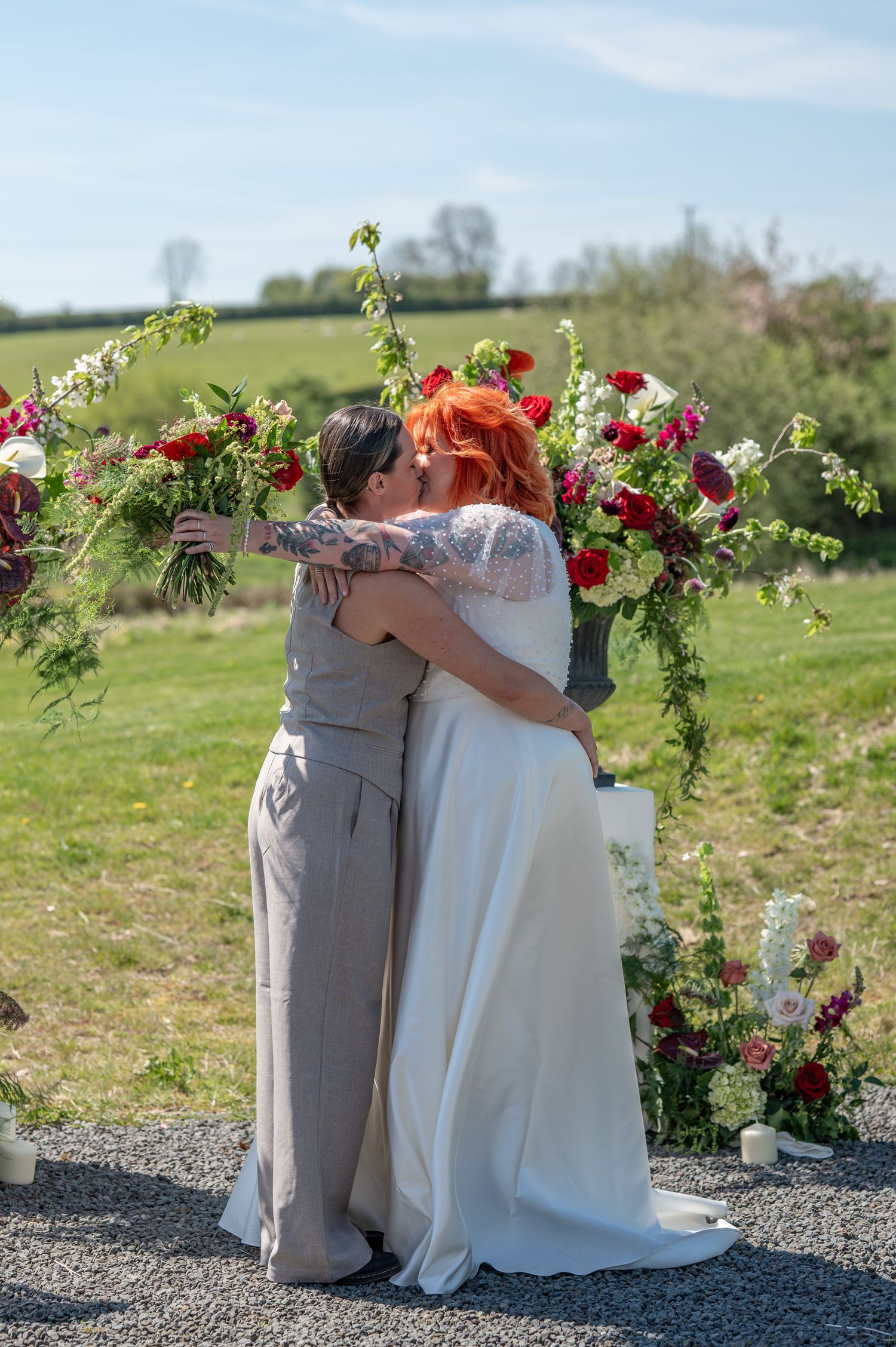 Two brides kiss in the sunshine at the Giraffe Shed, they are framed by beautiful flowers and there are fields in the background